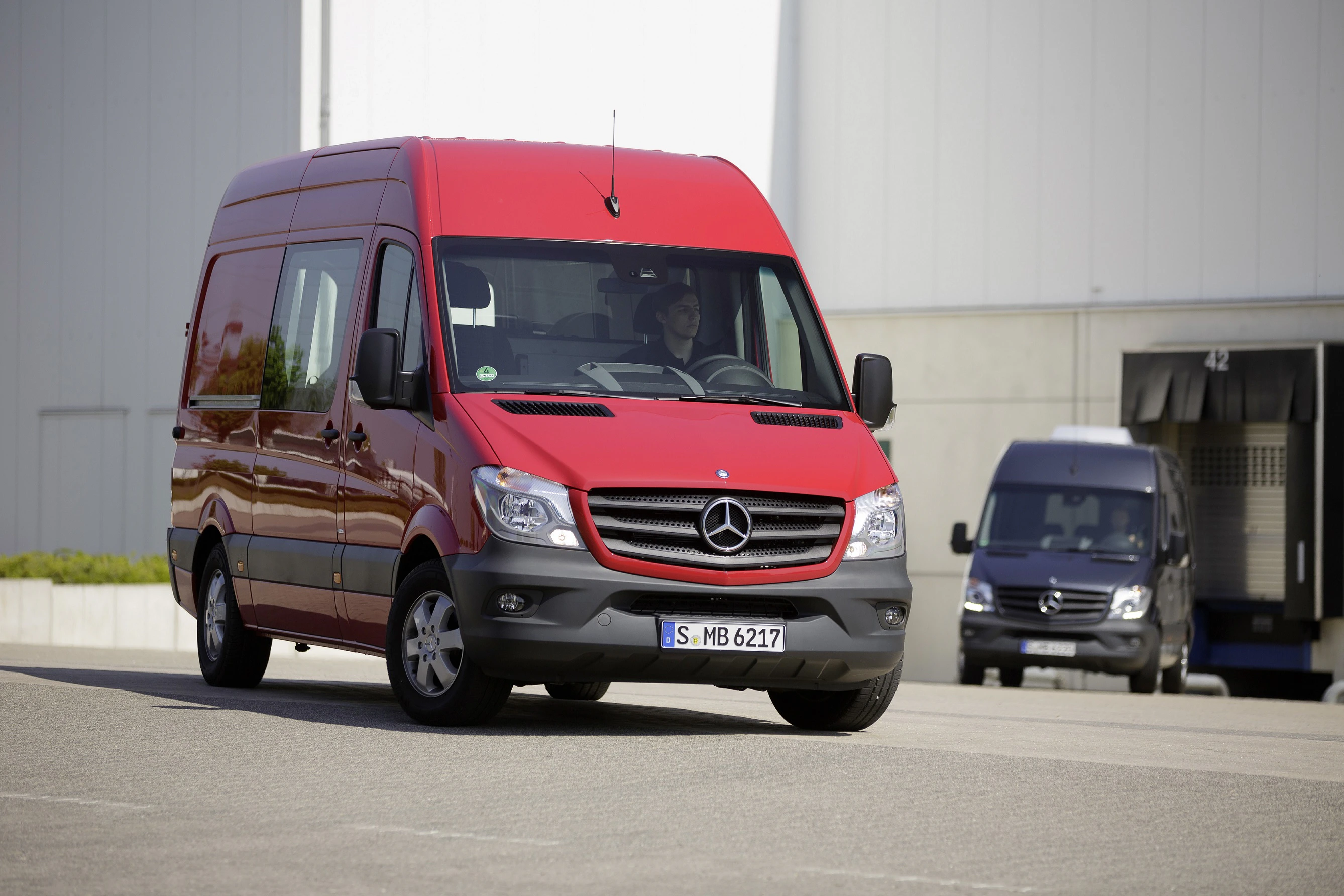 Roter Mercedes-Benz Sprinter fährt auf einem Industriehof. Im Hintergrund ist ein grauer Van und ein Gebäude zu sehen.