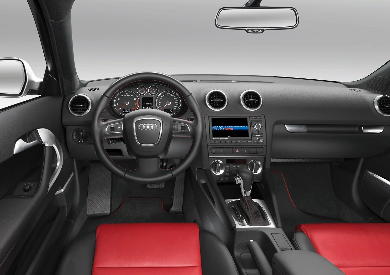 Innenraum eines Autos mit schwarzem Armaturenbrett, Lenkrad mit Audi-Logo, roten Sitzen und modernen Instrumenten.