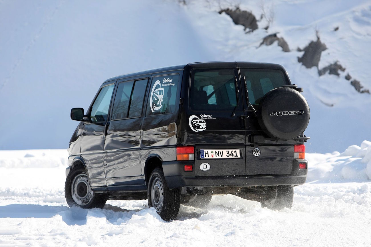 Ein schwarzer VW-Van fährt auf schneebedeckter Straße in bergiger Landschaft. Aufkleber und Reserverad am Heck des Fahrzeugs.