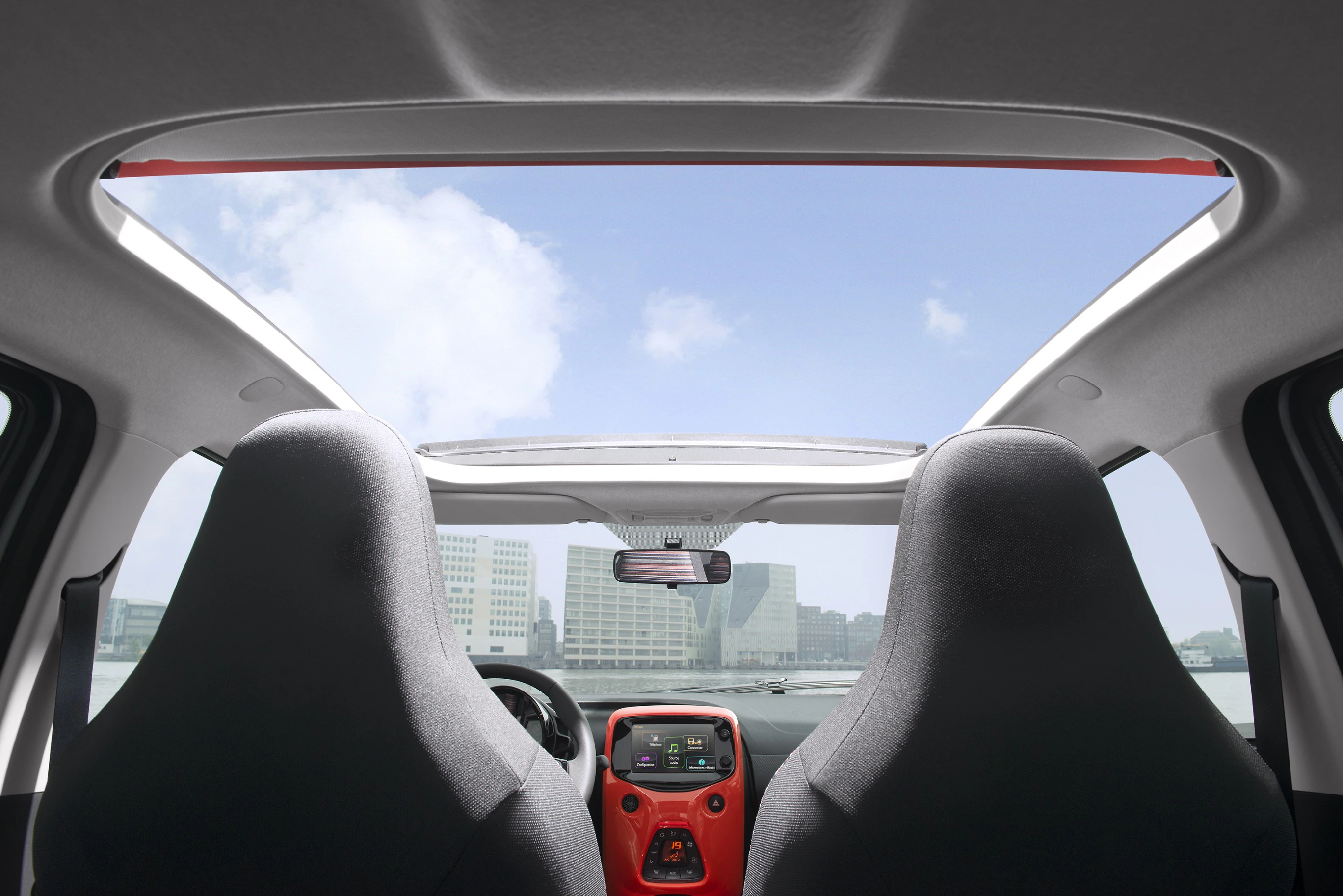 Innenraum eines Autos mit Panoramadach, Blick durch die Frontscheibe auf eine Stadtlandschaft im Hintergrund bei blauem Himmel.