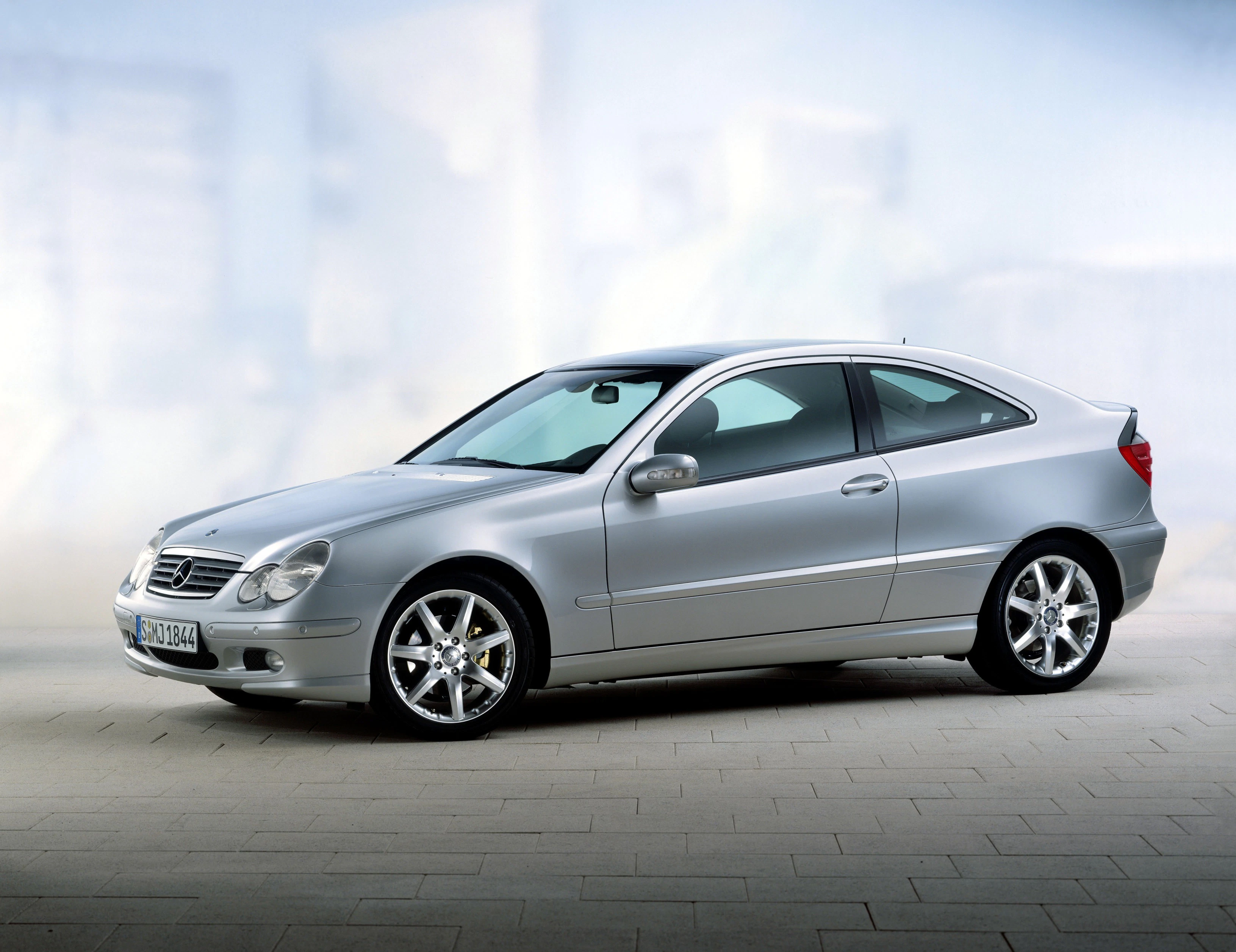 Silbernes Auto, Mercedes-Benz C-Klasse Coupé, auf gepflastertem Boden vor unscharfem, hellem Hintergrund, Seitenansicht.