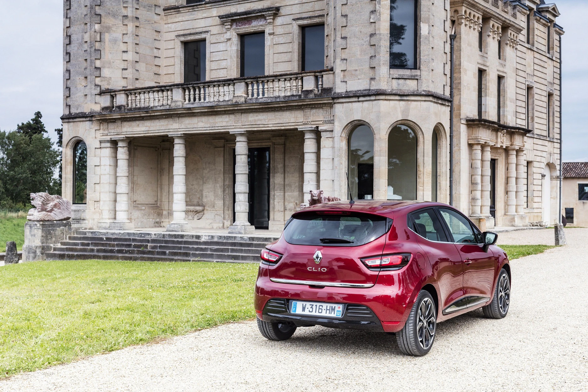 Ein roter Renault Clio parkt vor einem großen, alten Steingebäude mit Säulen und Rundbogenfenstern auf einem Kiesweg.