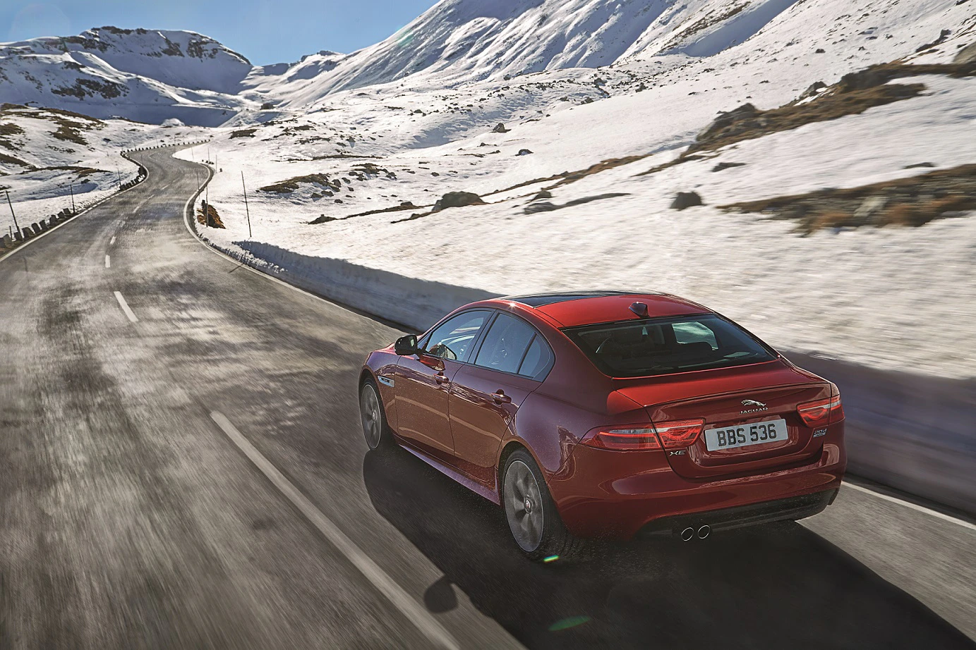 Rotes Auto fährt auf verschneiter Bergstraße bei klarem Himmel. Im Hintergrund sind schneebedeckte Hügel und eine kurvige Straße zu sehen.