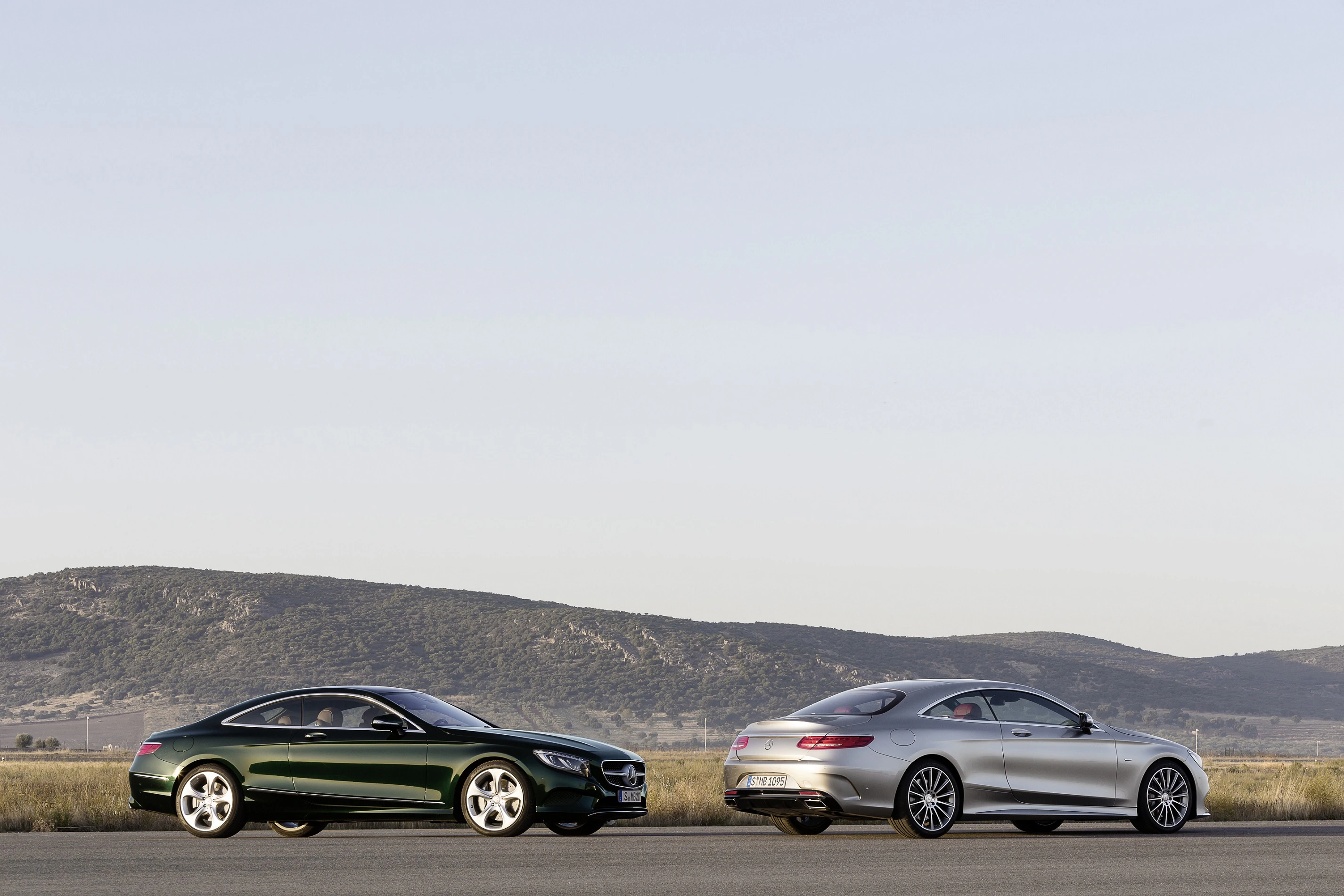 Zwei elegante Coupés auf einer freien Straße, umgeben von Hügeln und klarem Himmel. Das eine dunkelgrün, das andere silber.