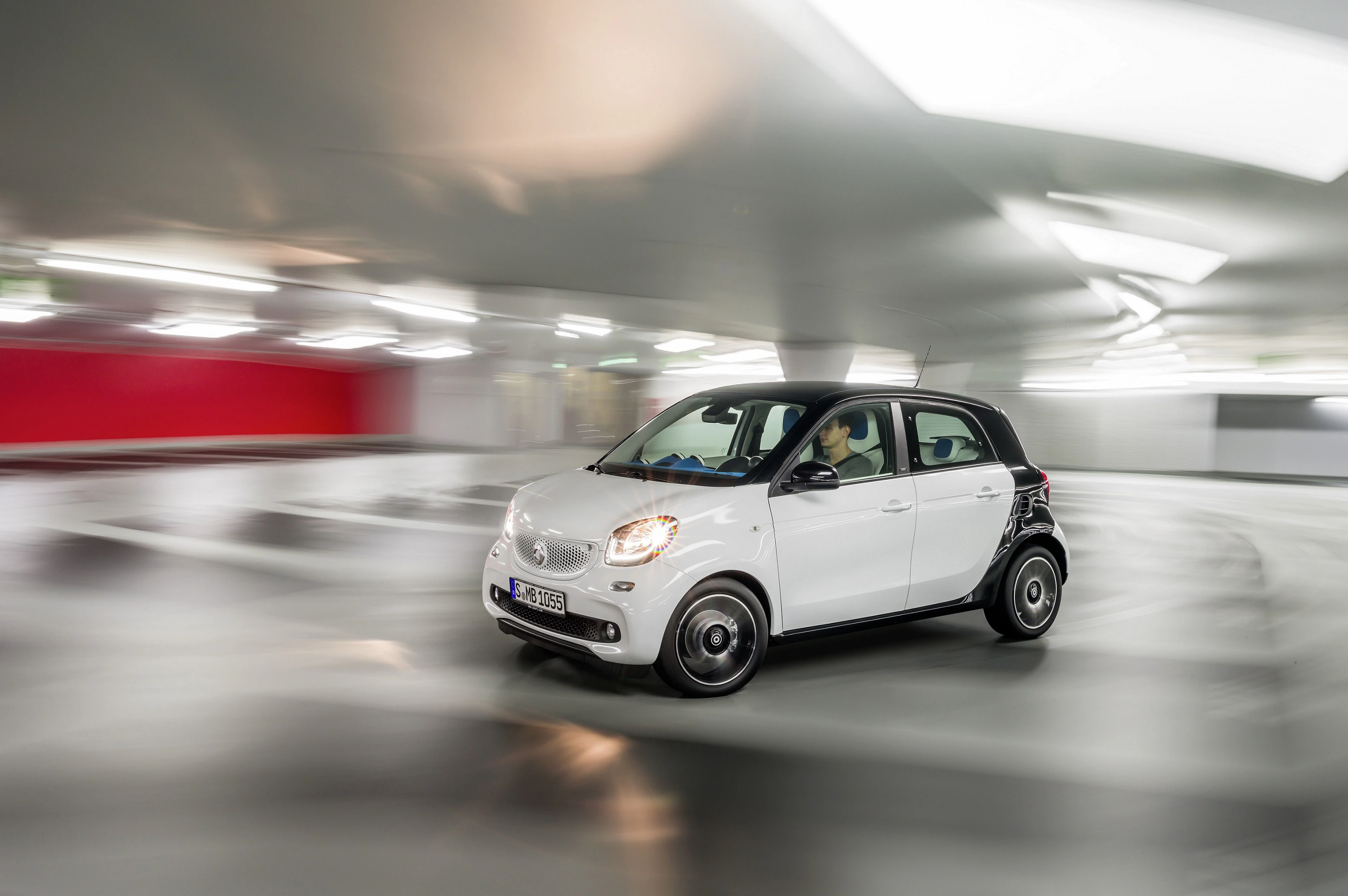 Weißer Kleinwagen fährt durch eine moderne, unscharf beleuchtete Parkgarage. Dynamische Aufnahme mit verschwommenem Hintergrund.