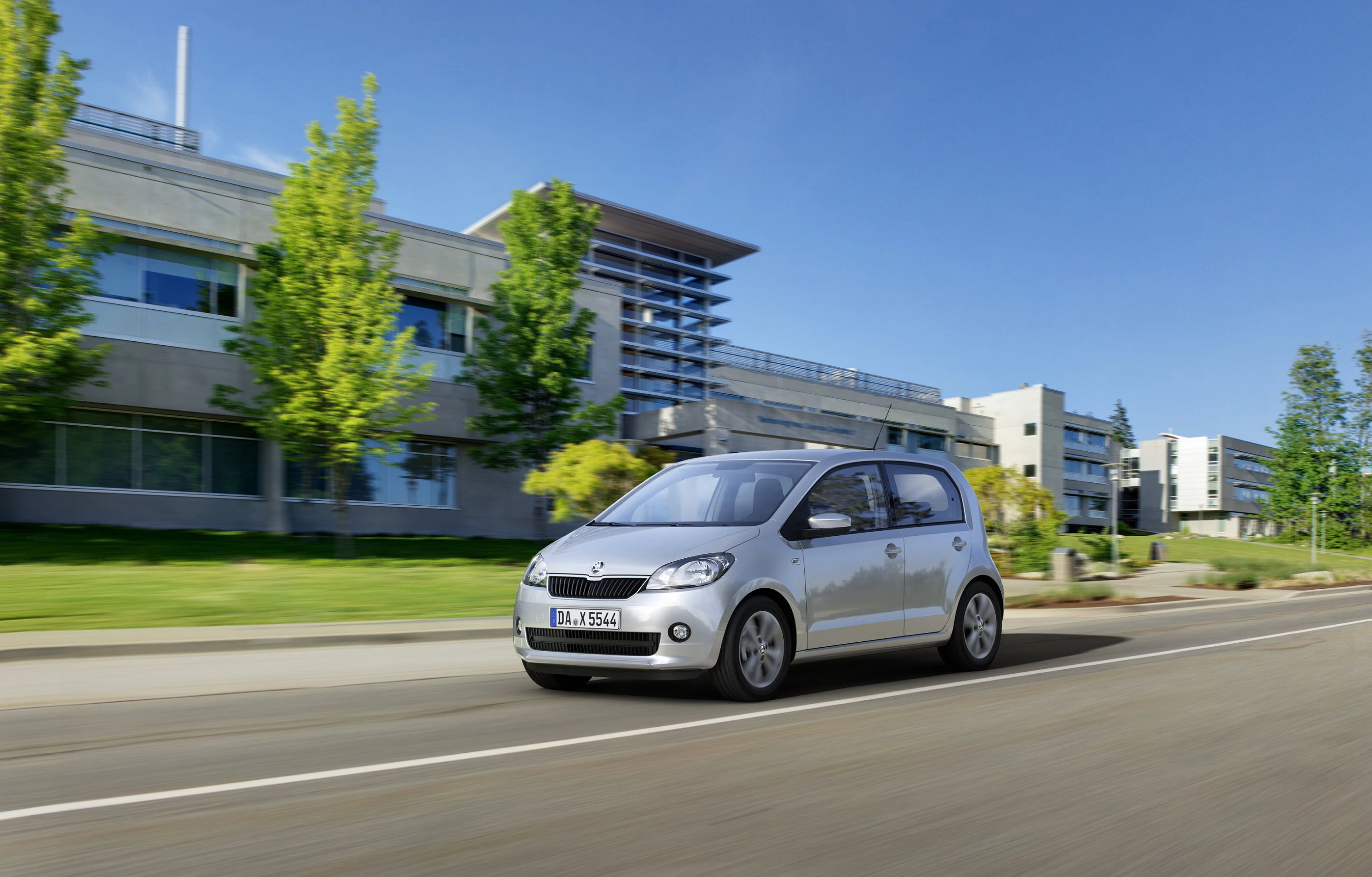 Silbernes Auto fährt auf einer Straße an modernen Gebäuden und grünen Bäumen vorbei bei klarem Himmel.