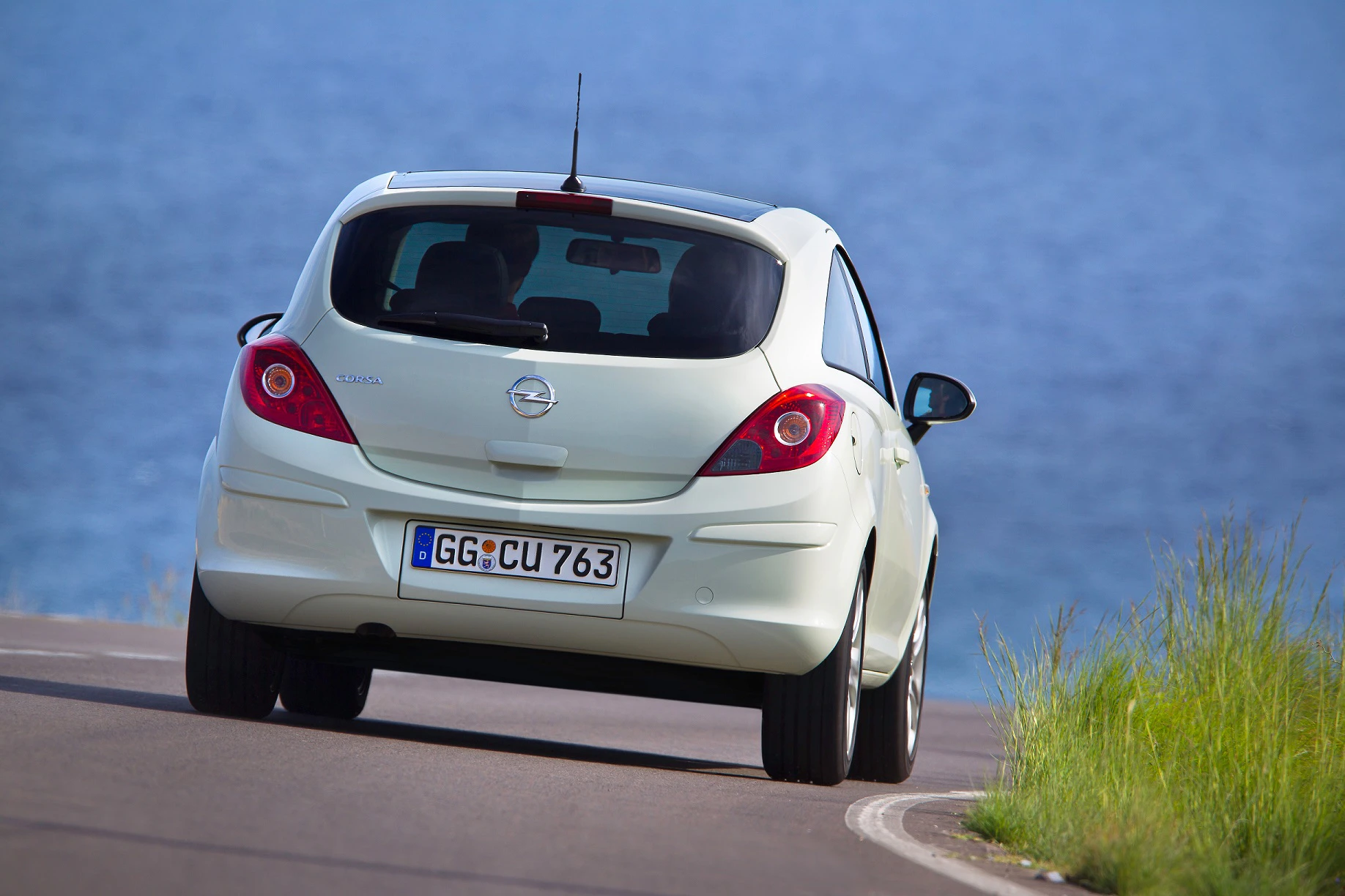 Ein cremefarbener Opel fährt auf einer Straße entlang der Küste mit blauem Meer im Hintergrund.
