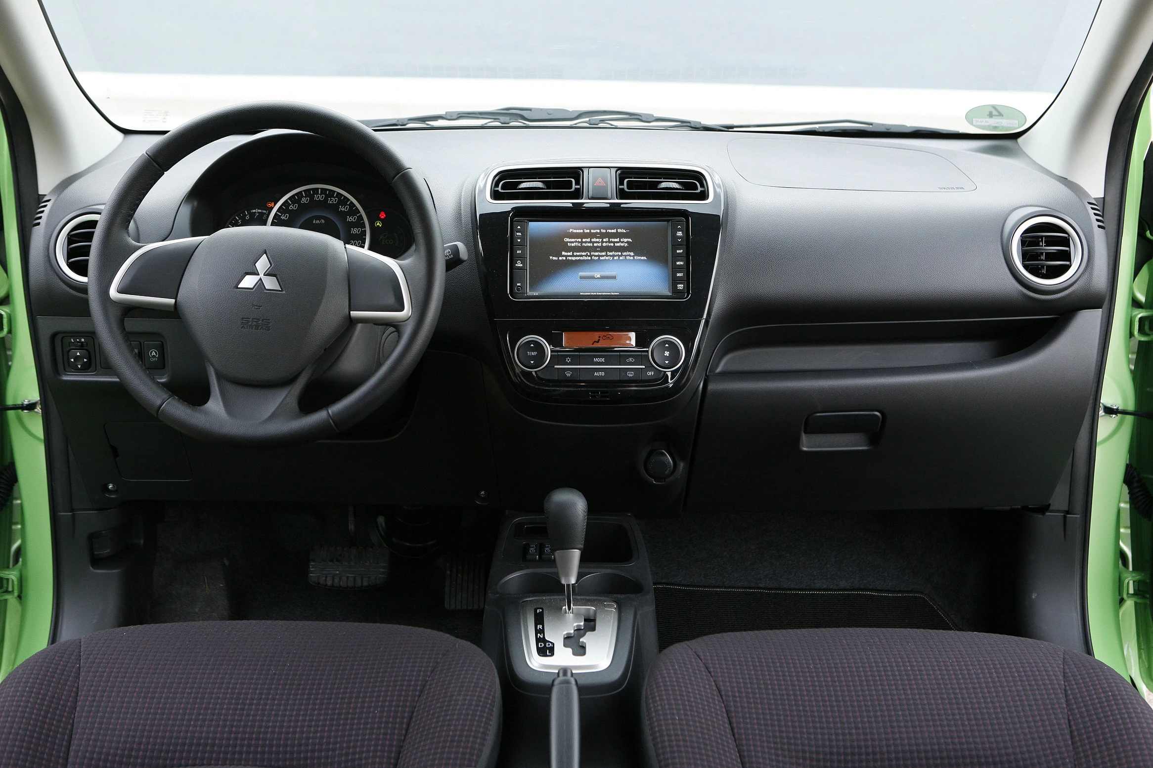 Armaturenbrett eines Autos mit Lenkrad links, zentralem Touchscreen, Lüftungsdüsen und Gangschaltung. Außenfarbe grün sichtbar.