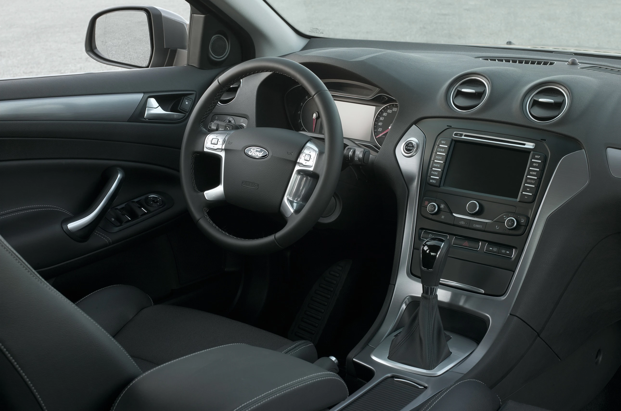 Innenraum eines Autos mit Lederlenkrad, zentralem Touchscreen und Armaturenbrett. Schwarze Sitze und Türverkleidungen sind sichtbar.