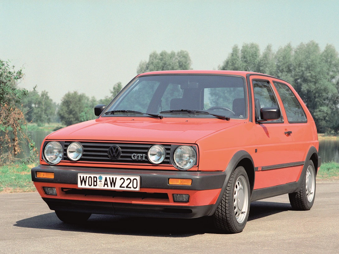Ein roter VW Golf II GTI steht auf einer Straße vor einer grünen Landschaft unter blauem Himmel, mit Bäumen im Hintergrund.