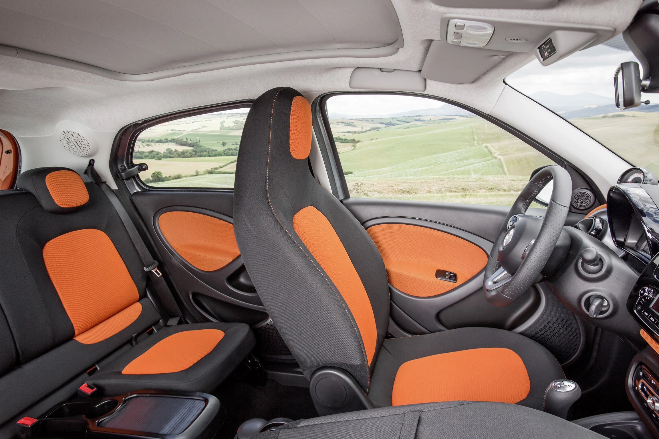 Innenraum eines Autos mit orange-schwarzen Sitzen und moderner Ausstattung, umgeben von einer Landschaft mit grünen Hügeln im Hintergrund.