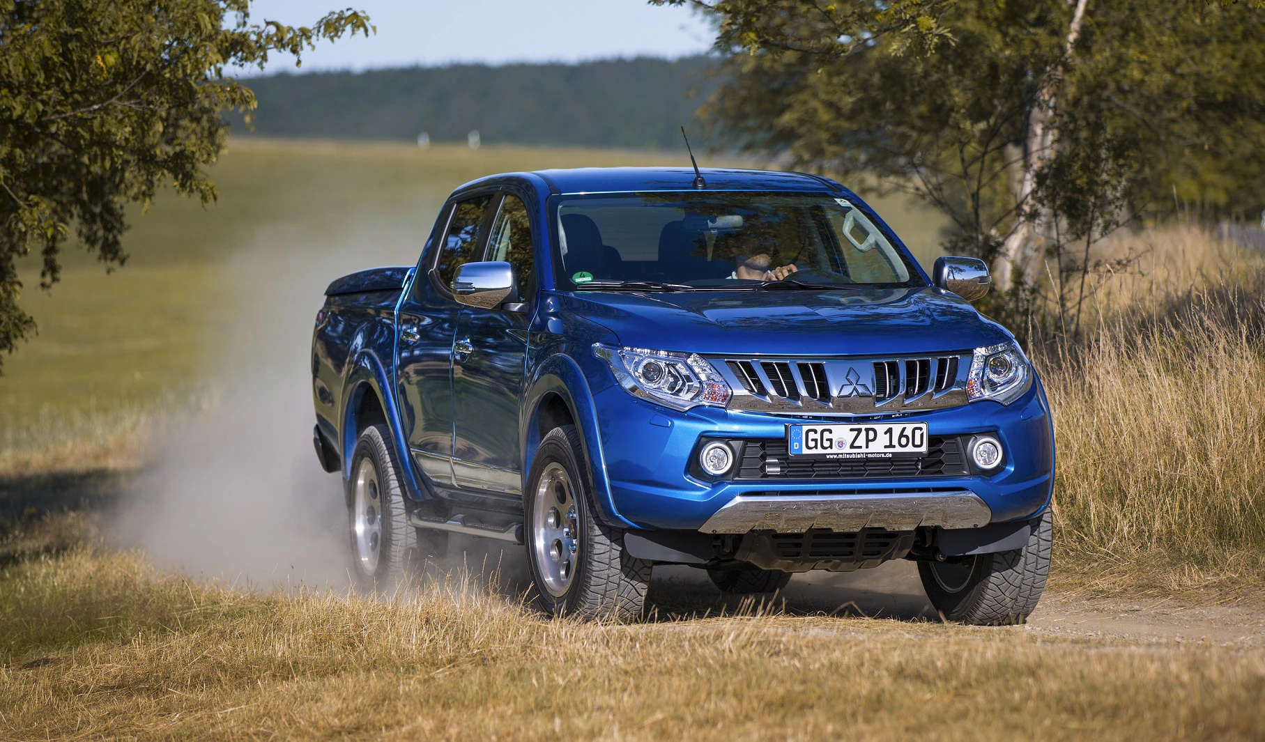 Blauer Pick-up-Truck fährt auf einem Feldweg durch eine ländliche Landschaft mit Bäumen, Staub wirbelt hinter den Reifen auf.
