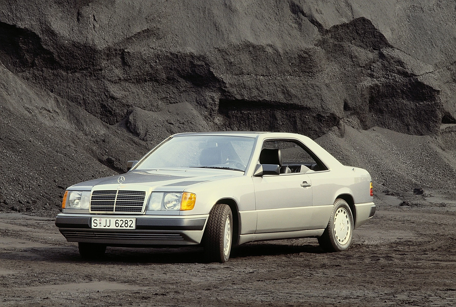 Ein grauer Mercedes-Benz Coupé der 1990er Jahre parkt vor einer dunklen, felsigen Kulisse.