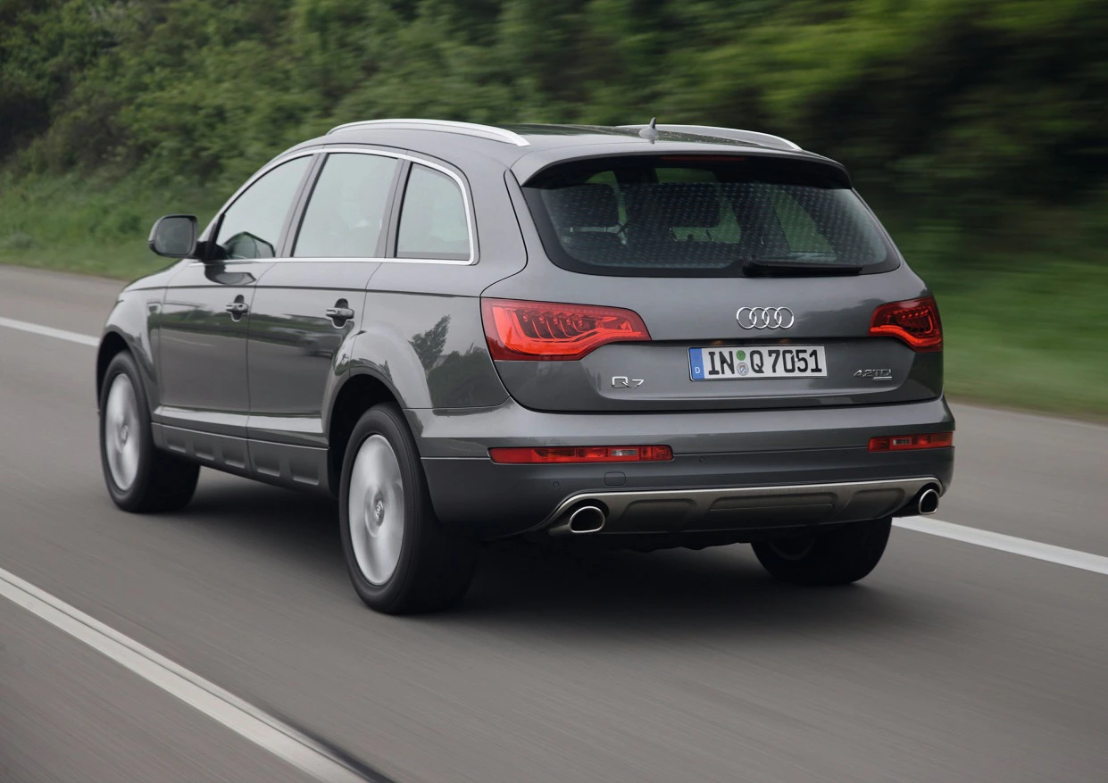 Grauer Audi Q7 fährt auf einer Landstraße. Rückansicht zeigt das Auto in Bewegung mit Bäumen im Hintergrund.