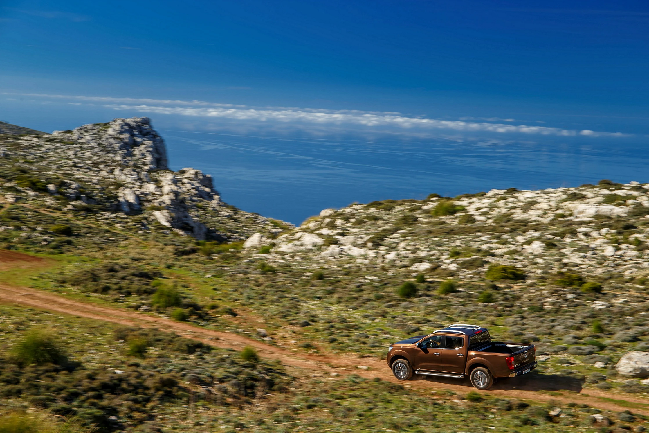 Ein brauner Pickup fährt auf einer unbefestigten Straße durch eine felsige Landschaft mit Meerblick und blauem Himmel im Hintergrund.