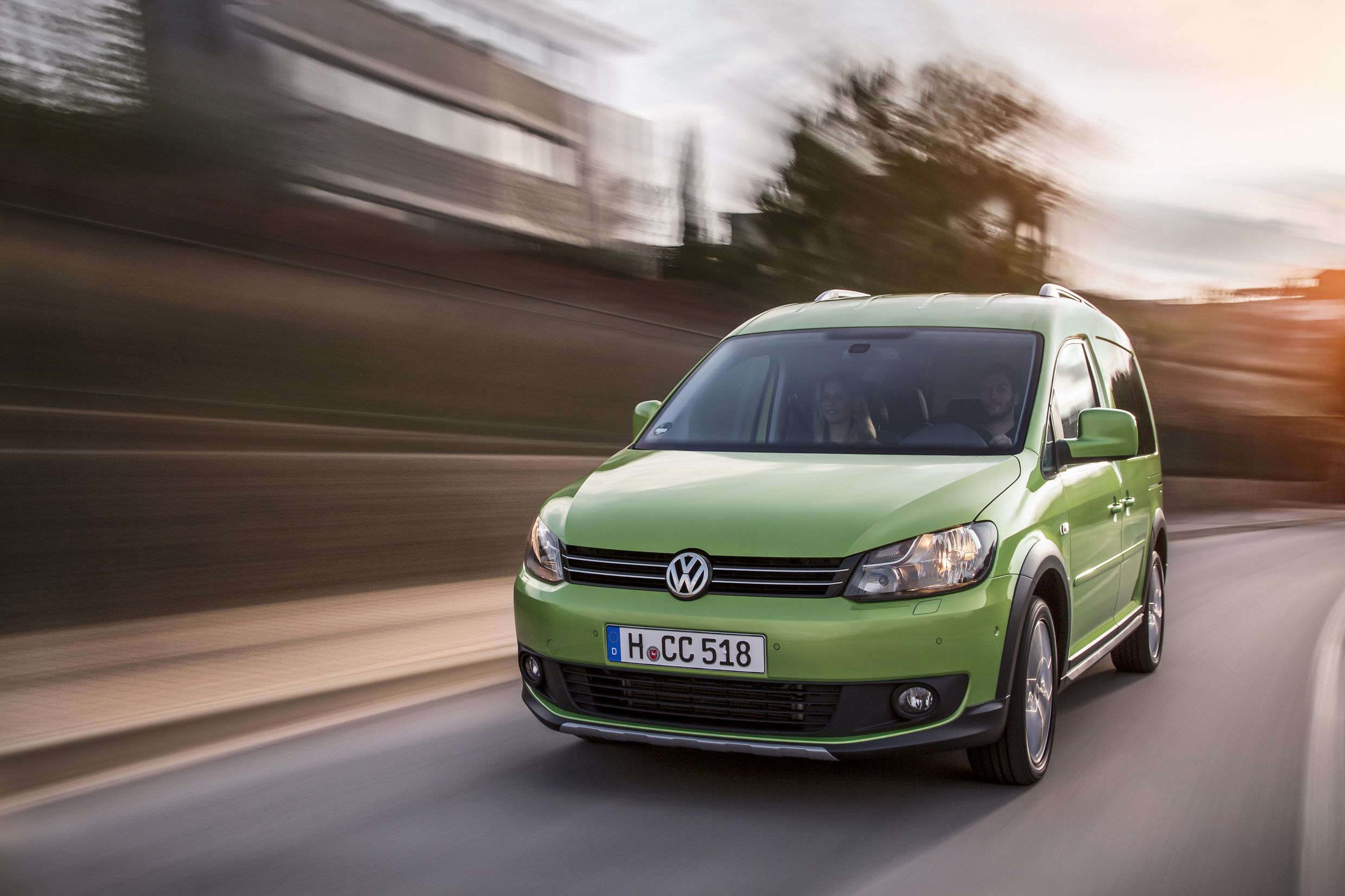 Grüner Volkswagen Caddy fährt auf einer Straße bei Sonnenuntergang, Gebäude im Hintergrund verschwommen und Geschwindigkeit suggerierend.