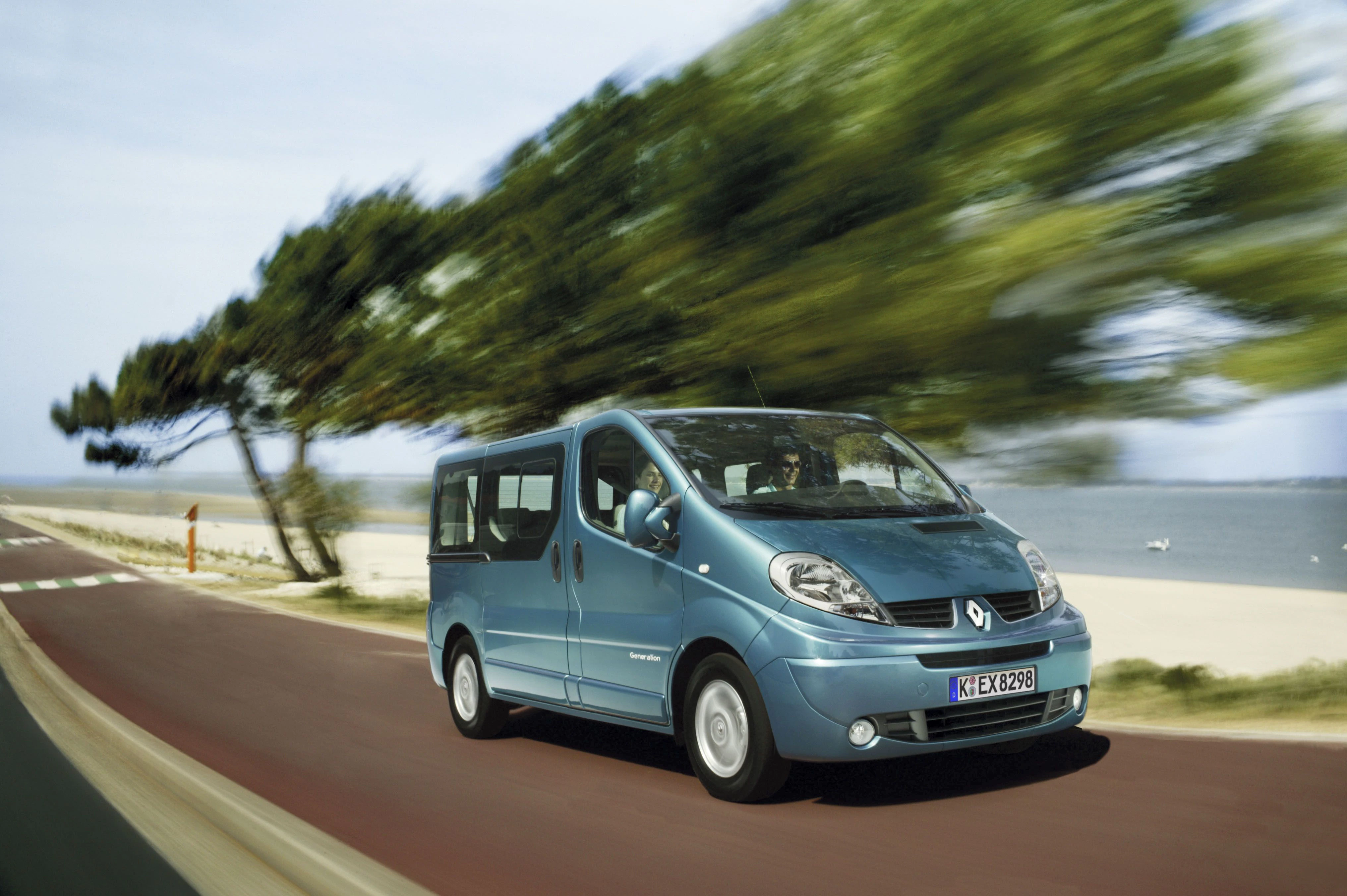 Blauer Renault Van fährt auf einer Küstenstraße vorbei an verschwommenen Bäumen. Meer im Hintergrund, sonniges Wetter.