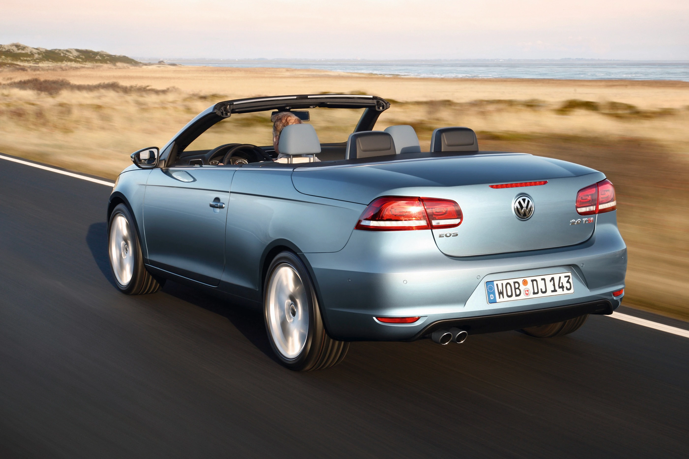 Blaues Volkswagen-Cabrio fährt auf einer Landstraße an einer Küstenlandschaft entlang, Verdeck geöffnet, sonnige Umgebung.