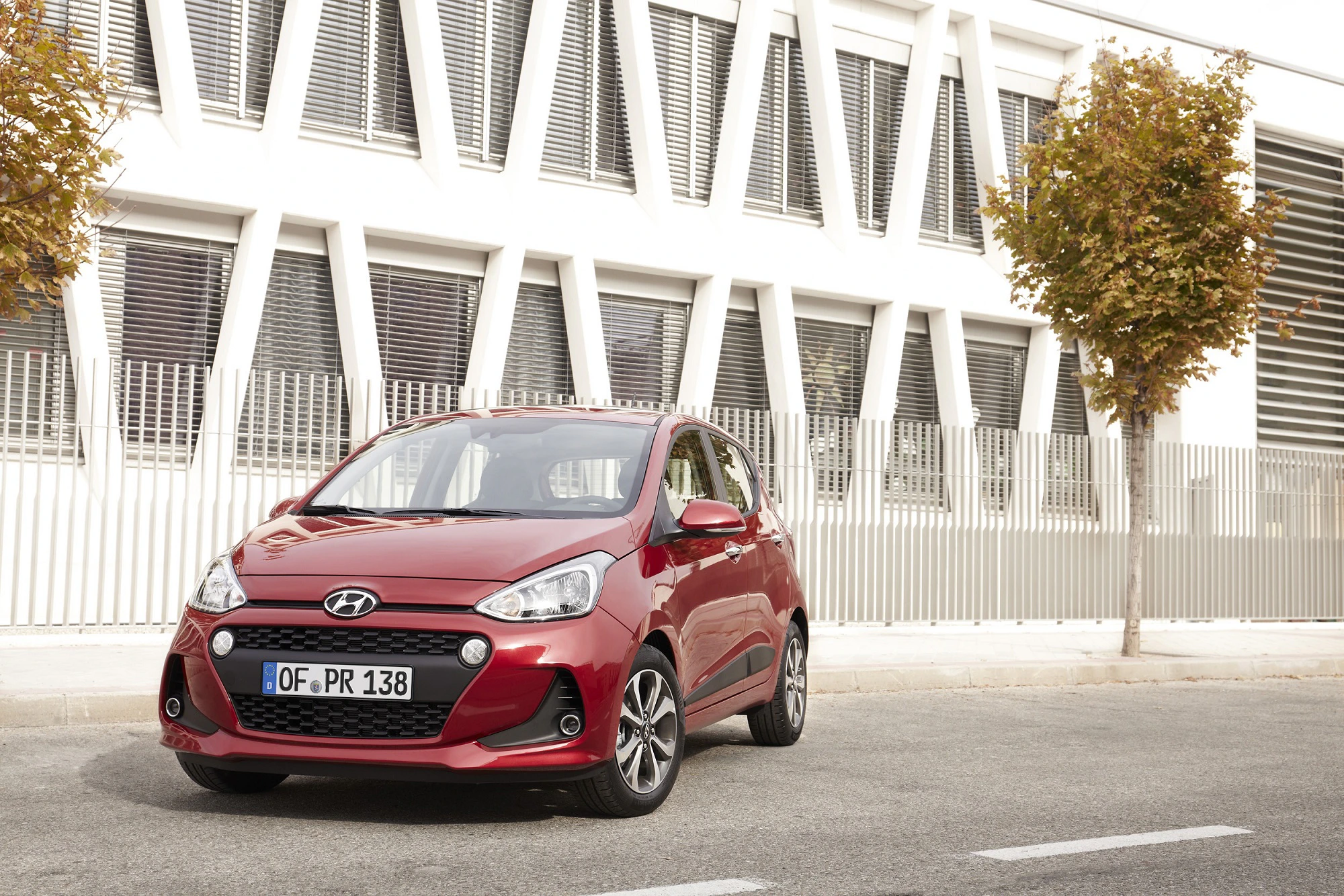 Roter Hyundai i10 parkt vor einem modernen Gebäude mit weißen Fensterläden und zwei kleinen Bäumen am Straßenrand.