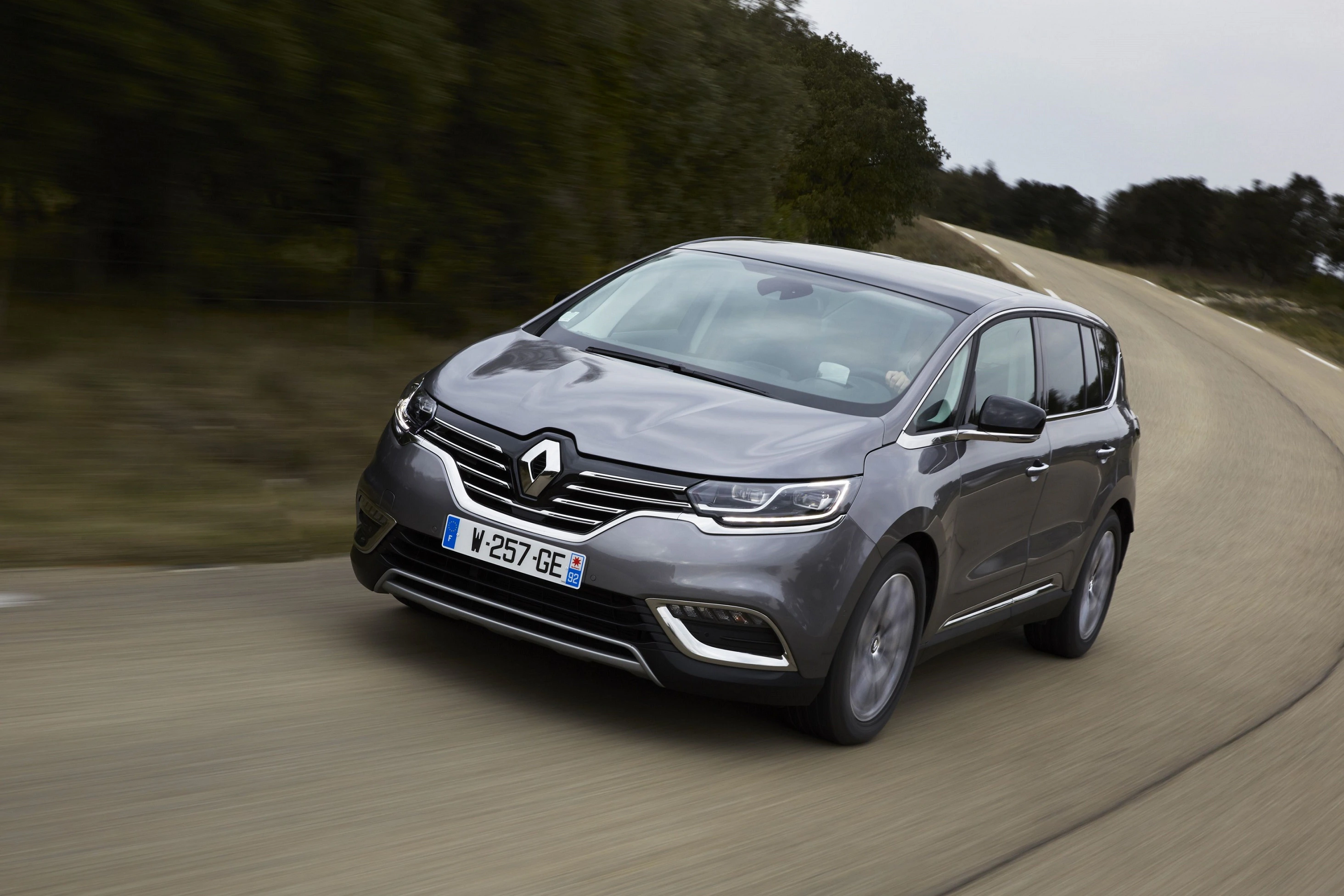 Grauer Renault Espace fährt auf kurviger Landstraße unter bewölktem Himmel. Bäume und Sträucher säumen die Straße.