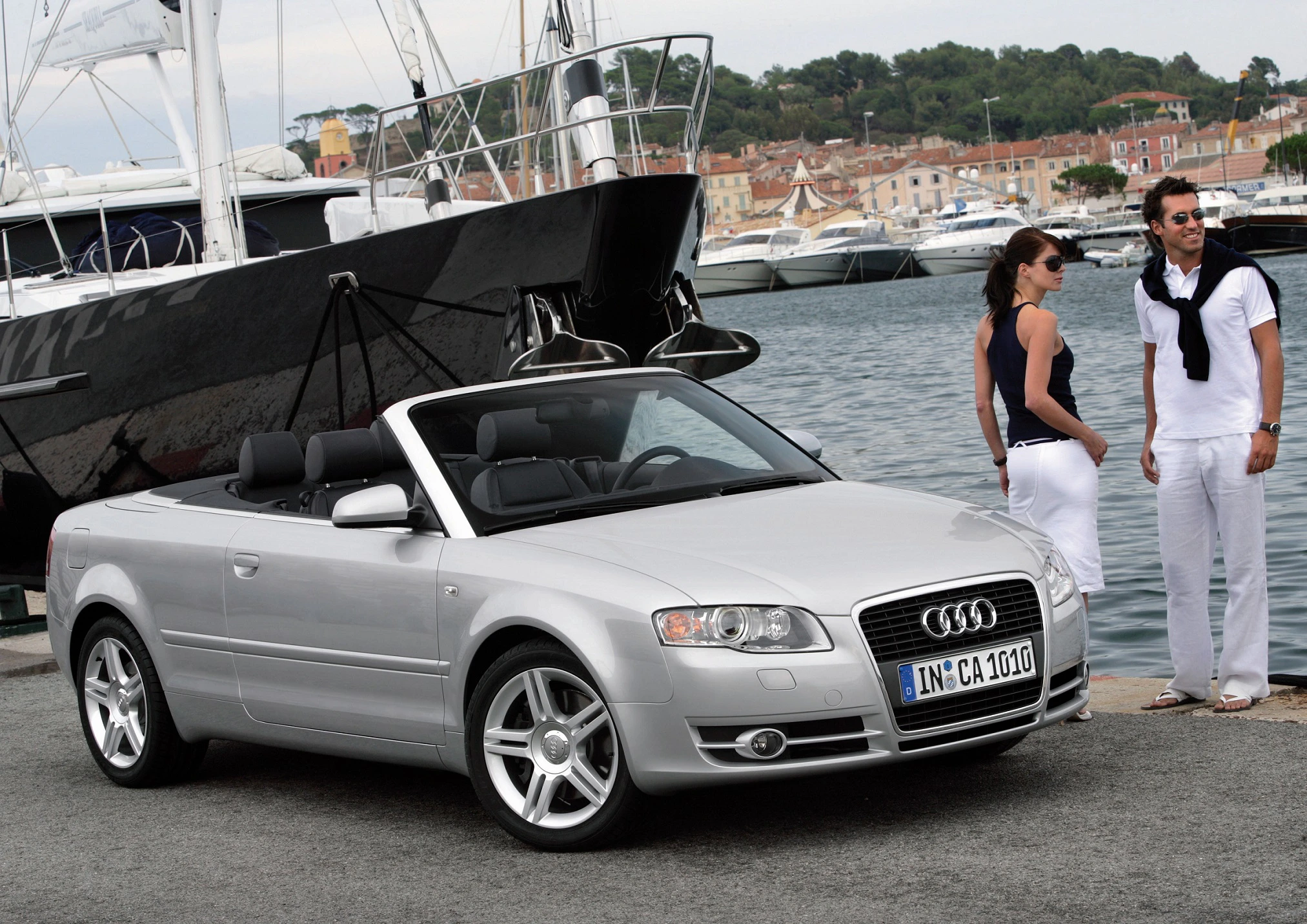 Silbernes Audi Cabriolet geparkt am Hafen, großes Segelboot im Hintergrund. Zwei Personen stehen entspannt am Kai.