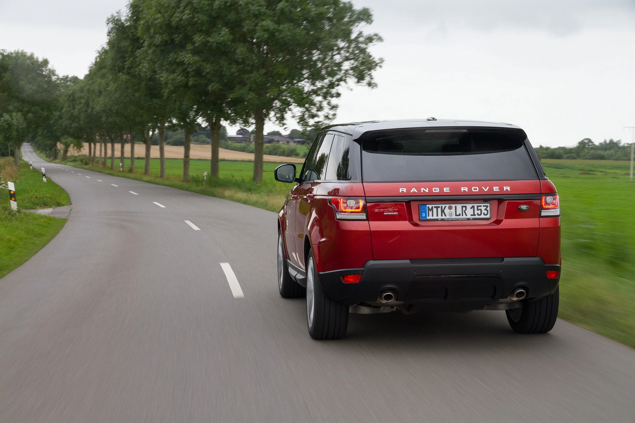 Roter Range Rover fährt auf einer Landstraße, umgeben von grünen Feldern und Bäumen.Nr.-Schild unscharf.