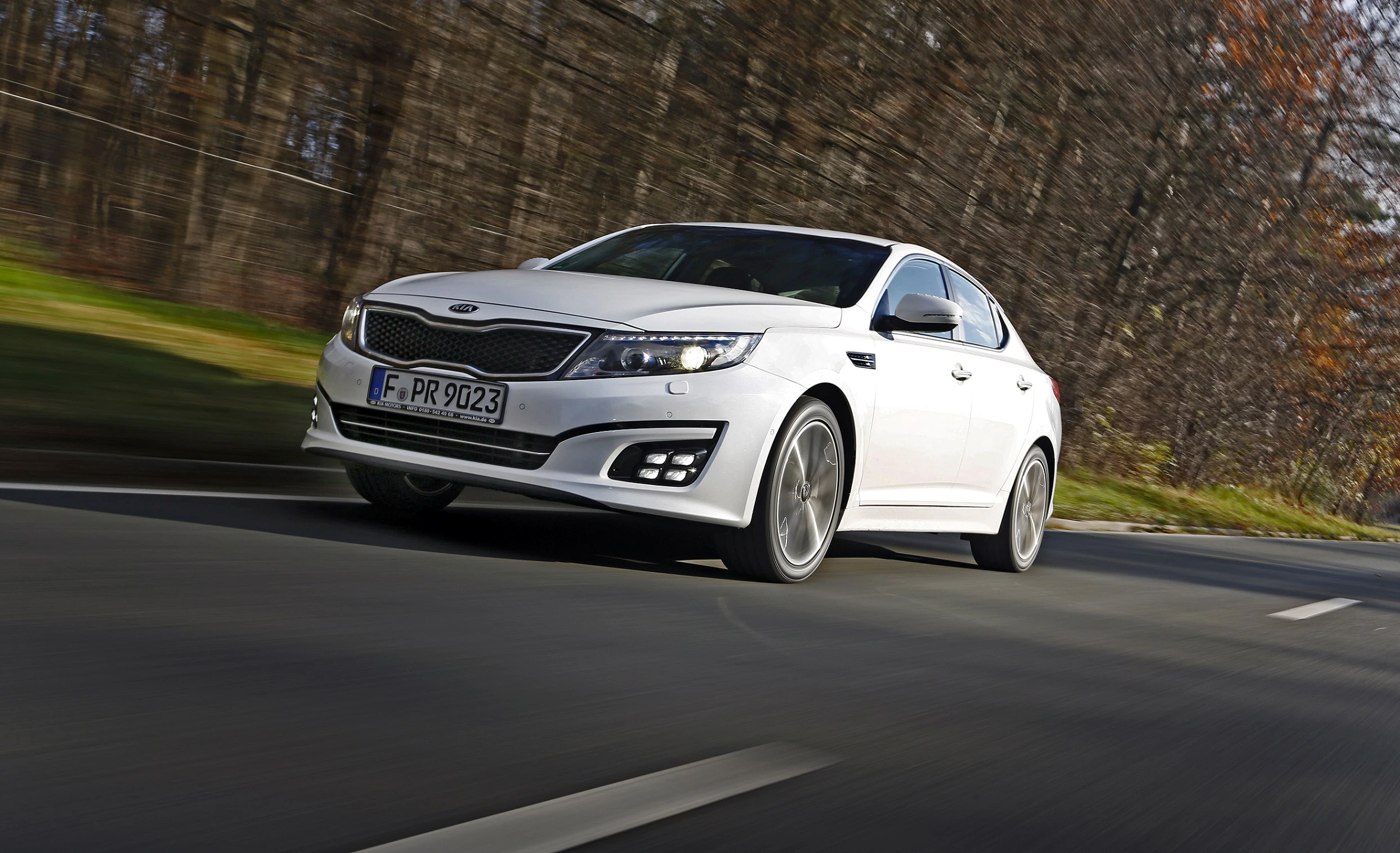 Weißer Kia Optima fährt schnell auf einer Landstraße durch eine herbstliche, bewaldete Umgebung.