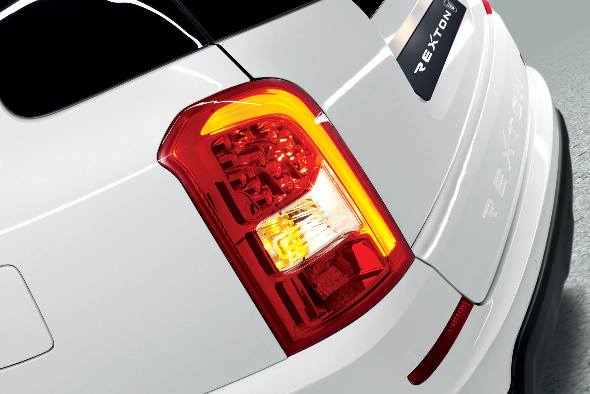 Rückansicht eines weißen Autos mit eingeschaltetem Rücklicht und reflektierender Oberfläche. Modellname "REXTON" am Fahrzeugheck sichtbar.