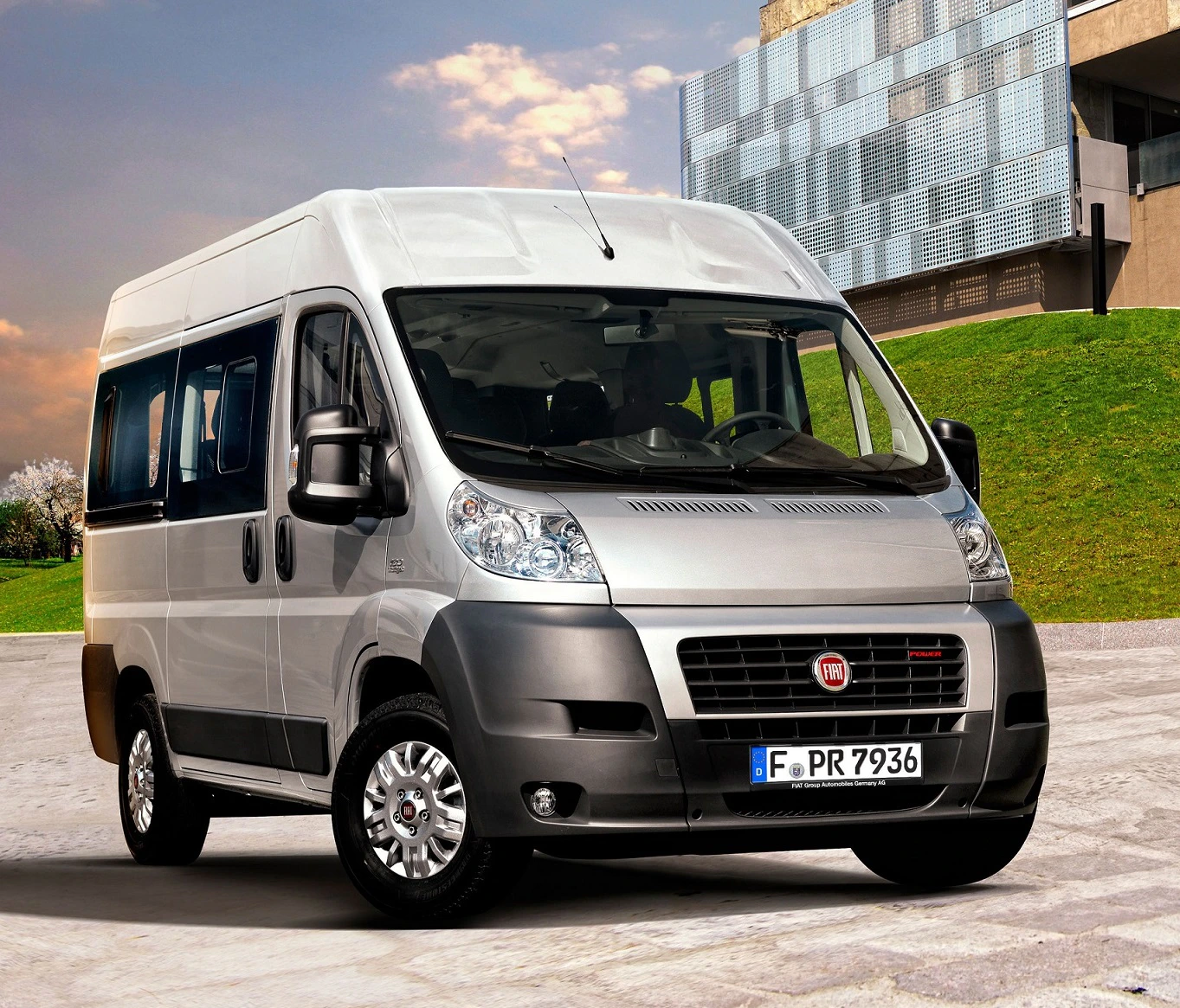 Silberner Fiat-Transporter parkt vor moderner Architektur und grünem Rasen unter blauem Himmel.
