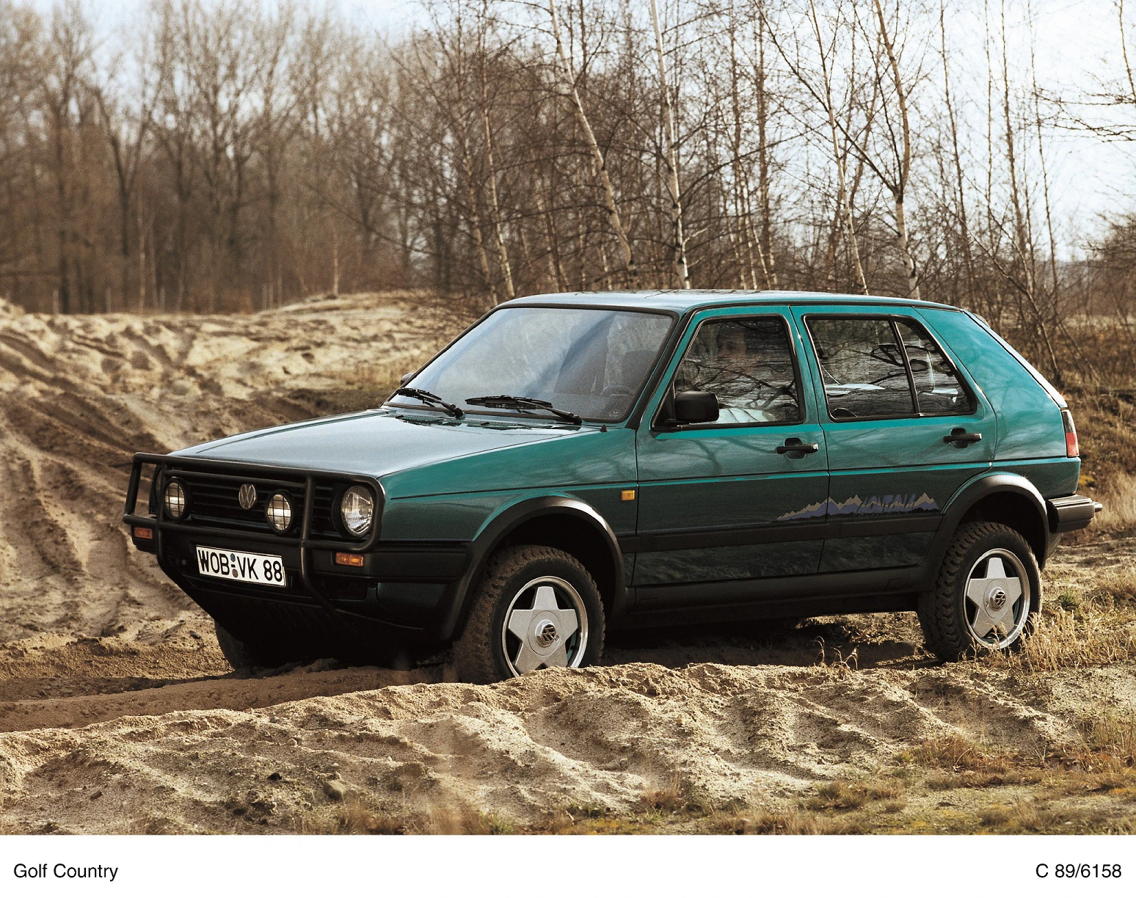 Grüner VW Golf Country fährt durch sandige, bewaldete Umgebung. Fahrzeug hebt sich durch Offroad-Reifen und hohe Bodenfreiheit hervor.