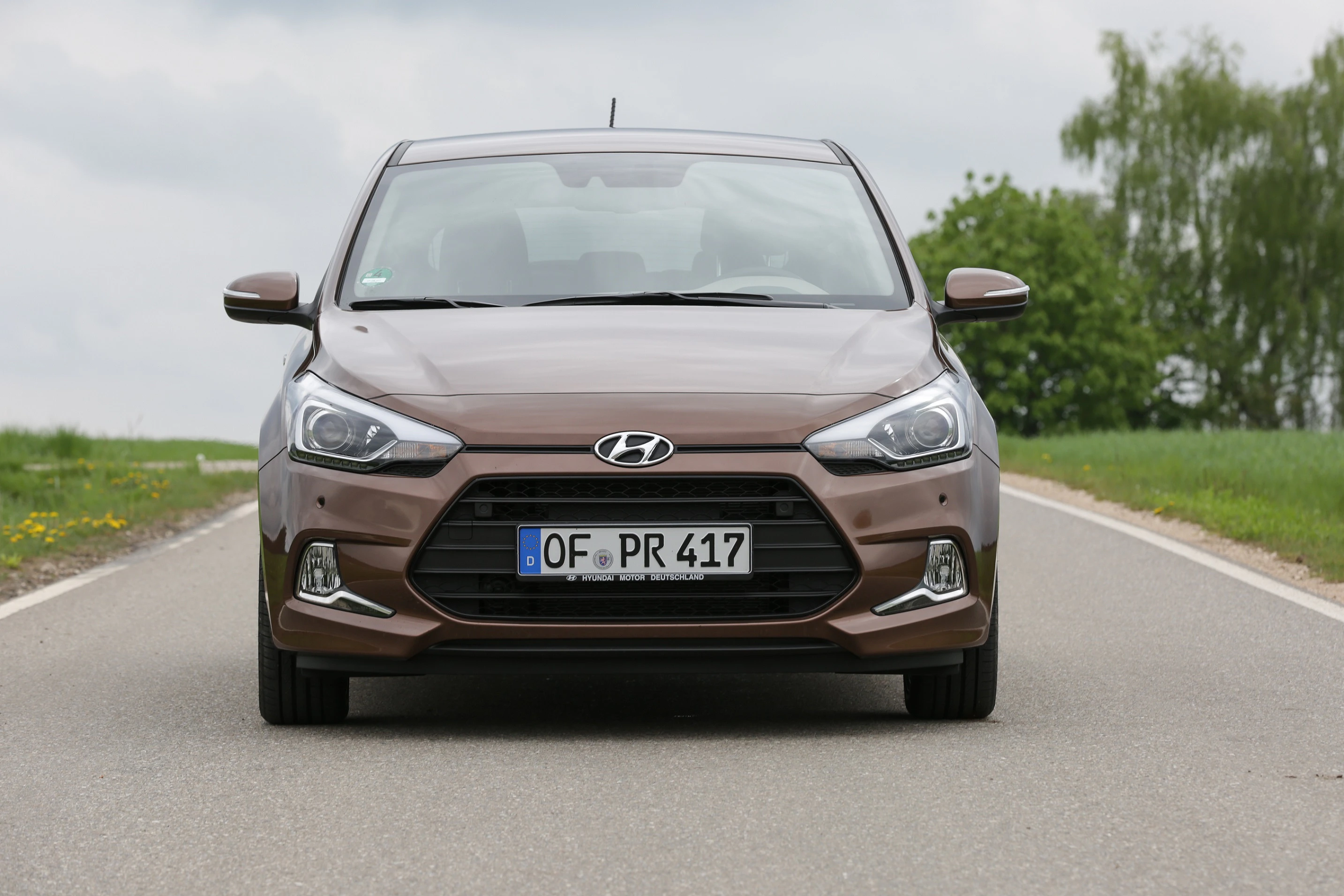 Brauner Hyundai i20 fährt auf ländlicher Straße. Kennzeichen: OF-PR 417. Bäume und Wiese im Hintergrund, bewölkter Himmel.