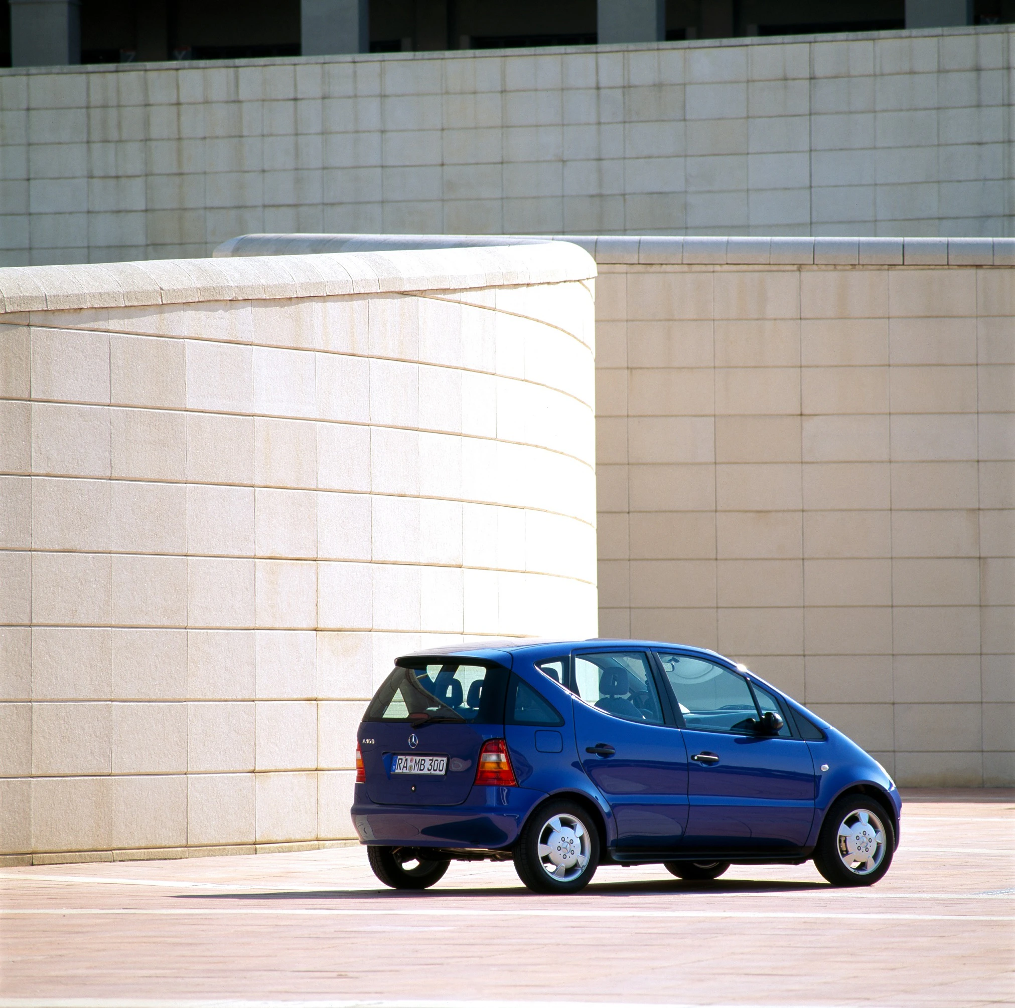 Blaues Auto parkt vor einer modernen, gewölbten Architektur aus beigen Steinen, bei hellem Tageslicht fotografiert.