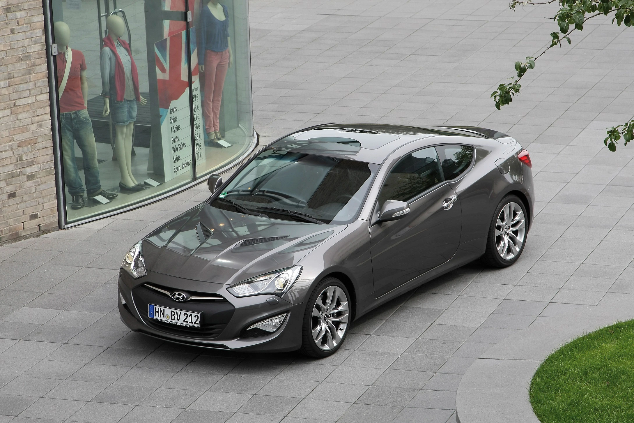 Graues Hyundai Coupé parkt auf gepflastertem Platz nahe Schaufenster mit Schaufensterpuppen.