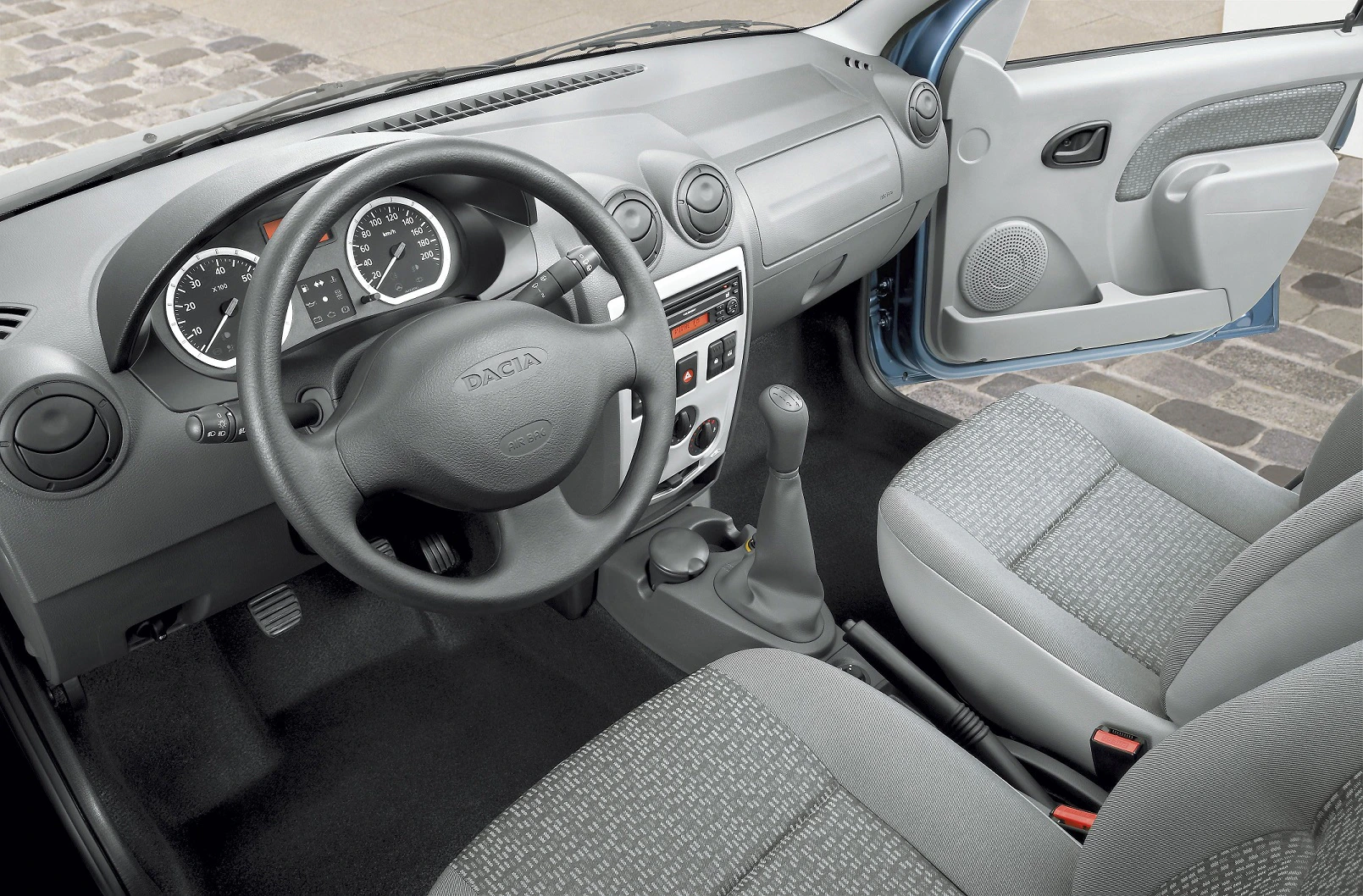 Innenraum eines Autos mit grauen Sitzen, Armaturenbrett mit Lenkrad, Schaltknüppel und geöffnetem Beifahrertür auf grauem Pflaster.