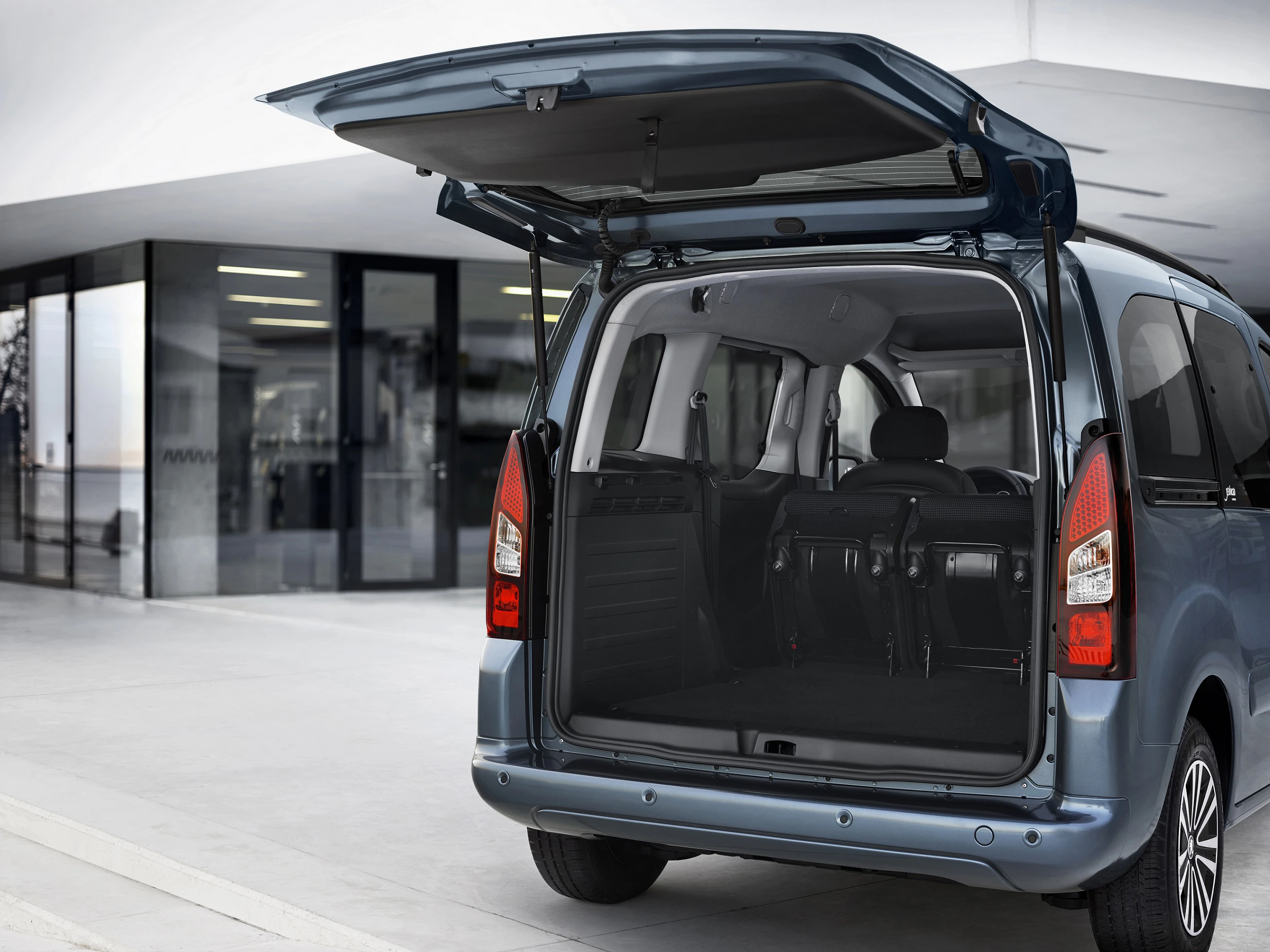 Ein blauer Minivan mit geöffneter Heckklappe steht vor einem modernen Gebäude. Der Innenraum ist leer und gut sichtbar.