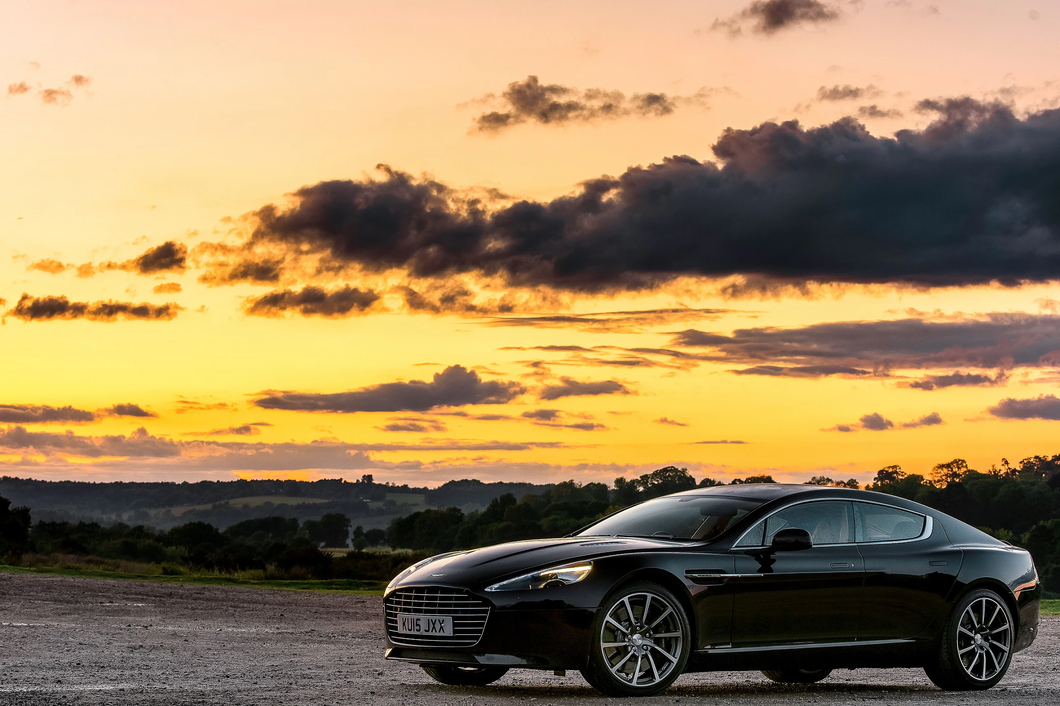 Ein schwarzes Auto steht in einer weiten Landschaft bei Sonnenuntergang. Der Himmel ist mit gelben und dunklen Wolken gefärbt.
