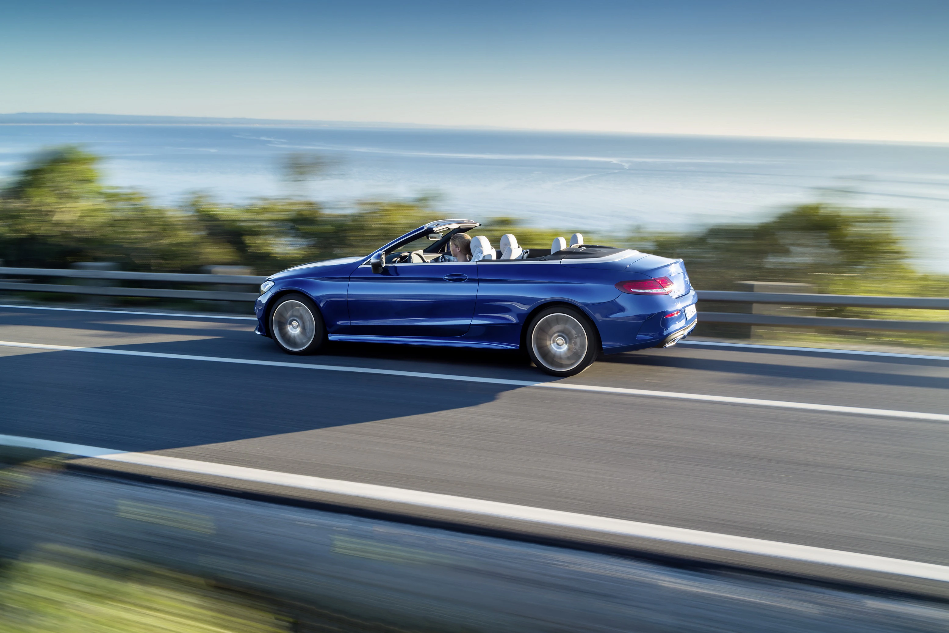 Blaues Cabriolet fährt schnell auf einer Küstenstraße, mit offenem Verdeck und Meer im Hintergrund.
