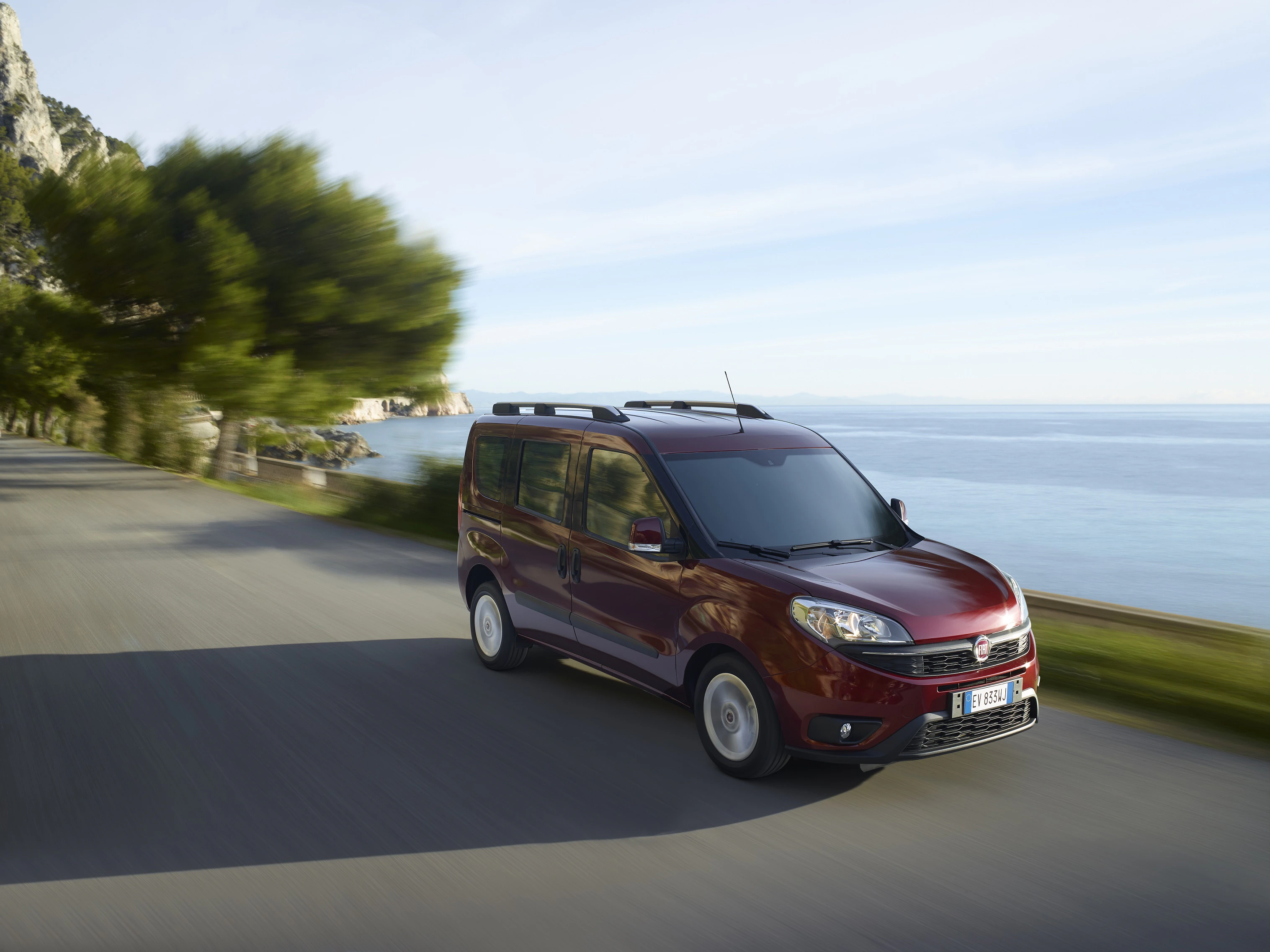 Roter Minivan fährt auf Küstenstraße mit Meerblick und bewaldetem Hintergrund bei sonnigem Wetter.