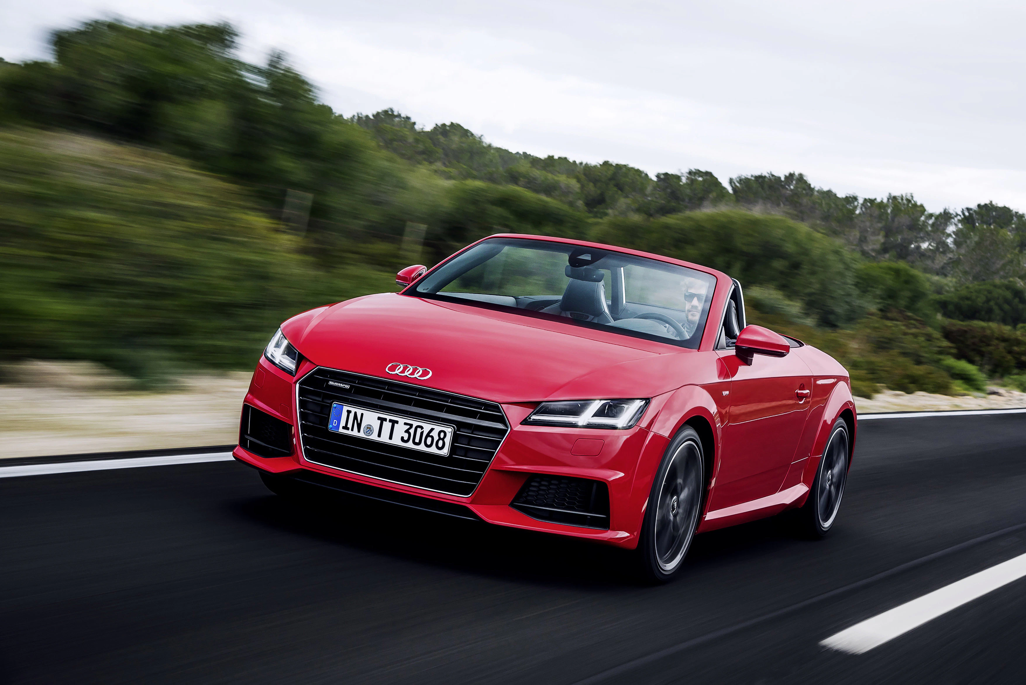 Roter Audi TT Cabrio fährt auf einer Straße, umgeben von grüner Landschaft und bewölktem Himmel. Dynamische und elegante Fahrzeugdarstellung.