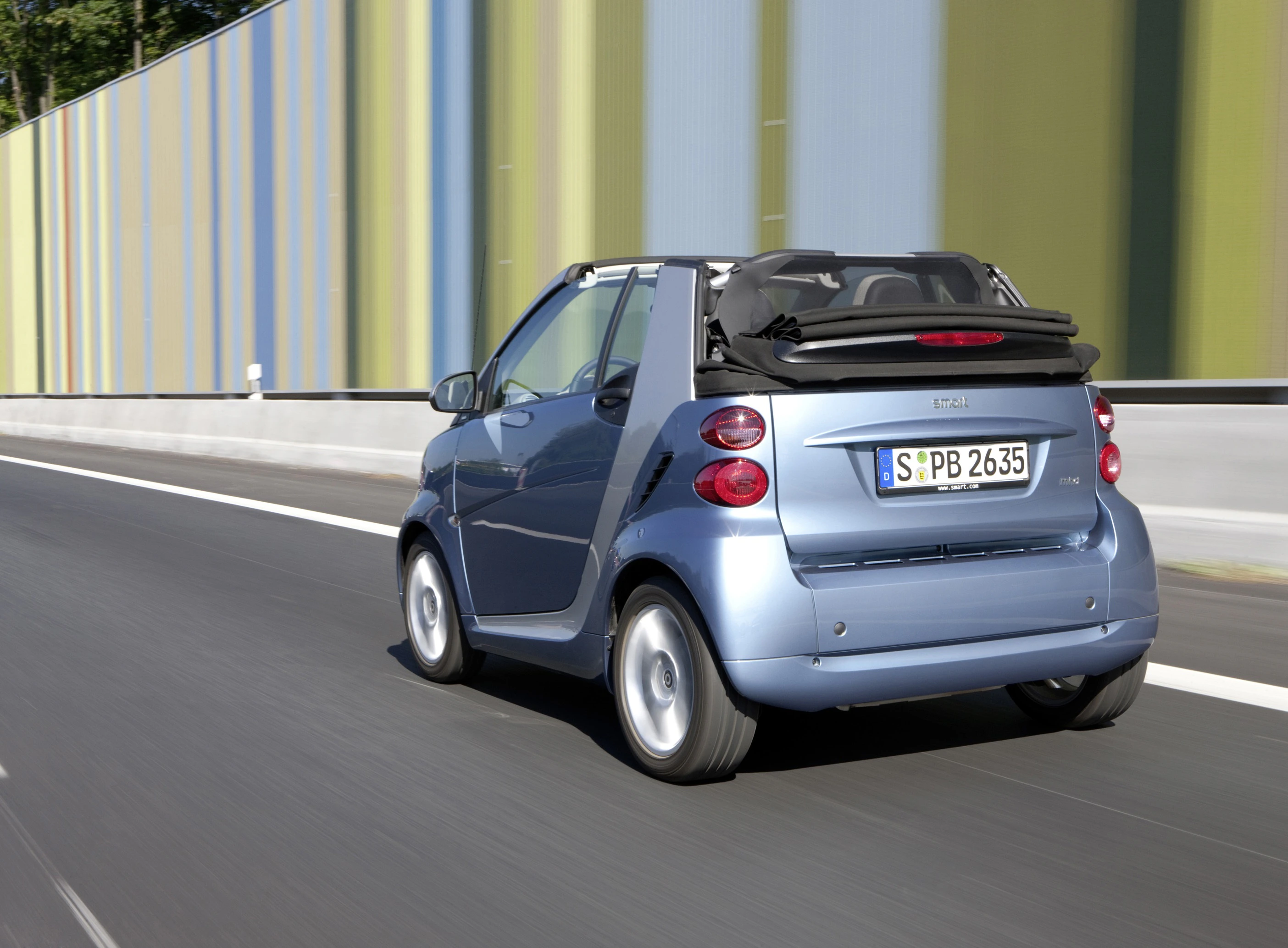 Ein blauer Smart Cabrio fährt auf einer Straße vor bunten Schallschutzwänden. Das Fahrzeug hat ein geöffnetes Verdeck.