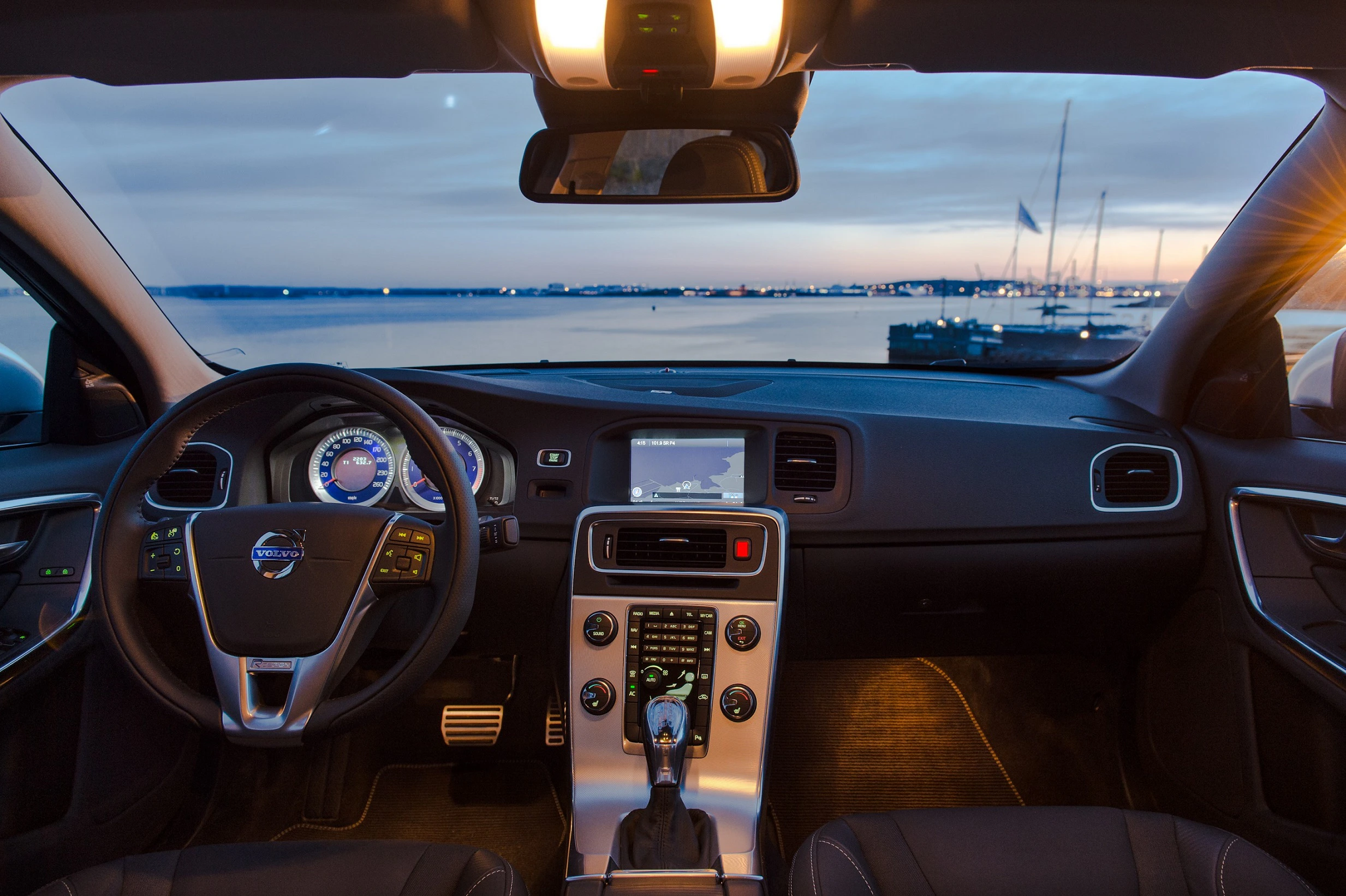 Innenansicht eines Autos mit Blick auf das Meer bei Sonnenuntergang. Lenkrad, Armaturenbrett und Bildschirm im Vordergrund.