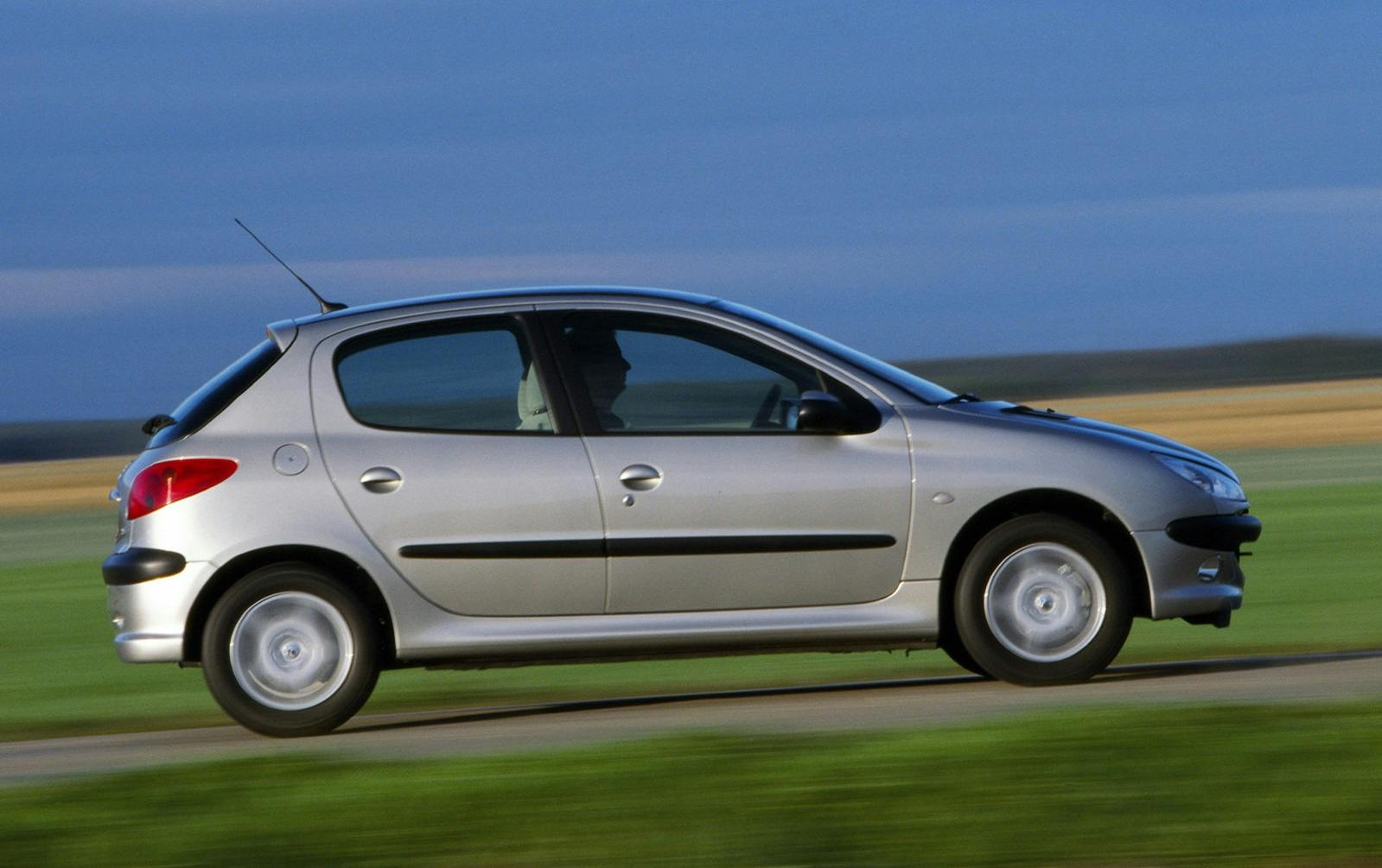 Silberner Kleinwagen fährt schnell auf einer Landstraße, unscharfer Hintergrund aus Feldern und blauem Himmel.
