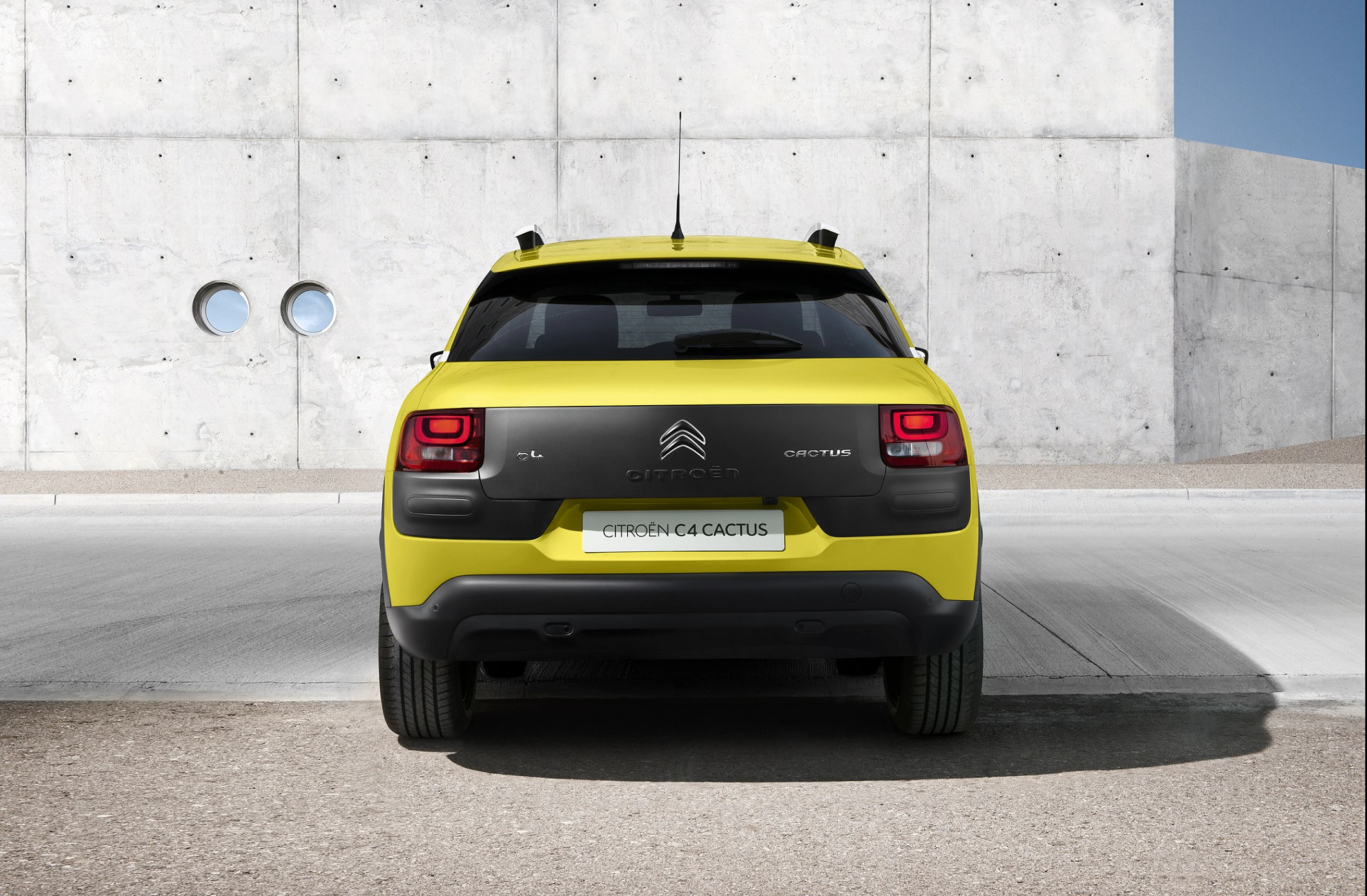 Gelber Citroën C4 Cactus von hinten vor einer grauen Betonwand mit zwei runden Fenstern, geparkt auf einer geteerten Fläche.