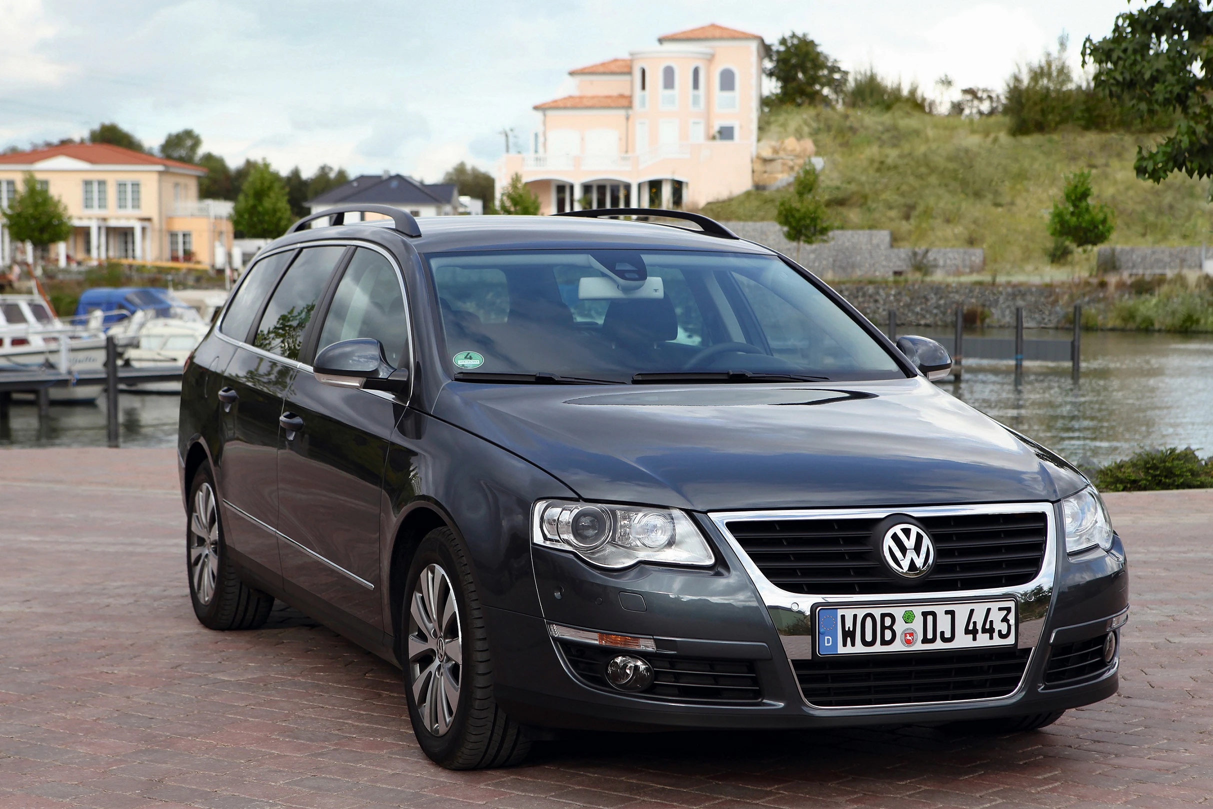 Dunkelgrauer Volkswagen Kombi parkt vor Wasser und Häusern. Nummernschild sichtbar. Pflastersteinstraße im Vordergrund.