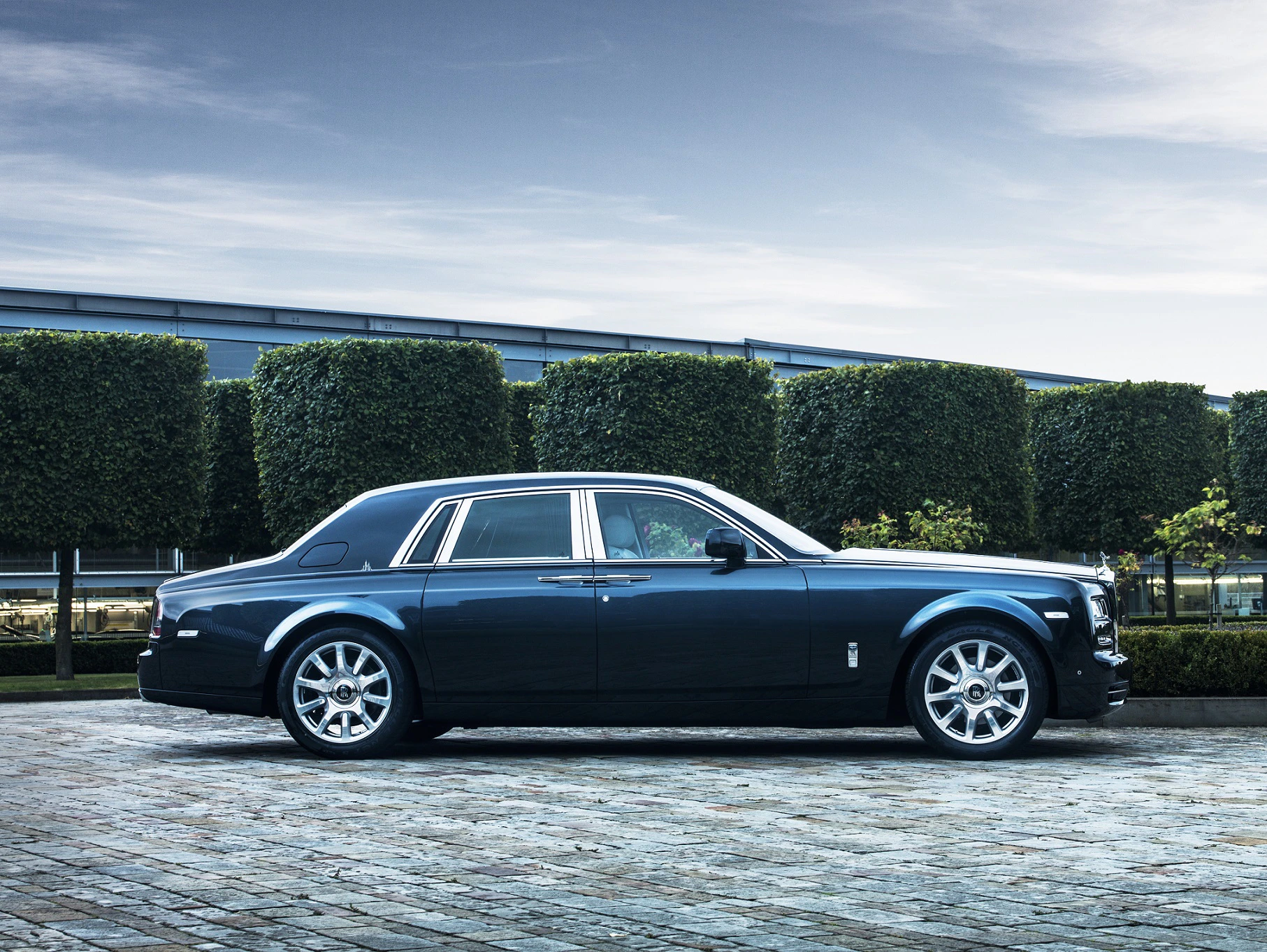 Luxuriöser dunkelblauer Rolls-Royce Phantom, seitlich auf Kopfsteinpflaster, mit geschnittenen Büschen und modernem Gebäude im Hintergrund.