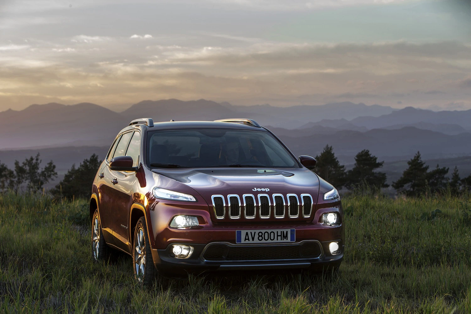 Roter Jeep-SUV steht auf einer Wiese vor einer Berglandschaft bei Sonnenuntergang.