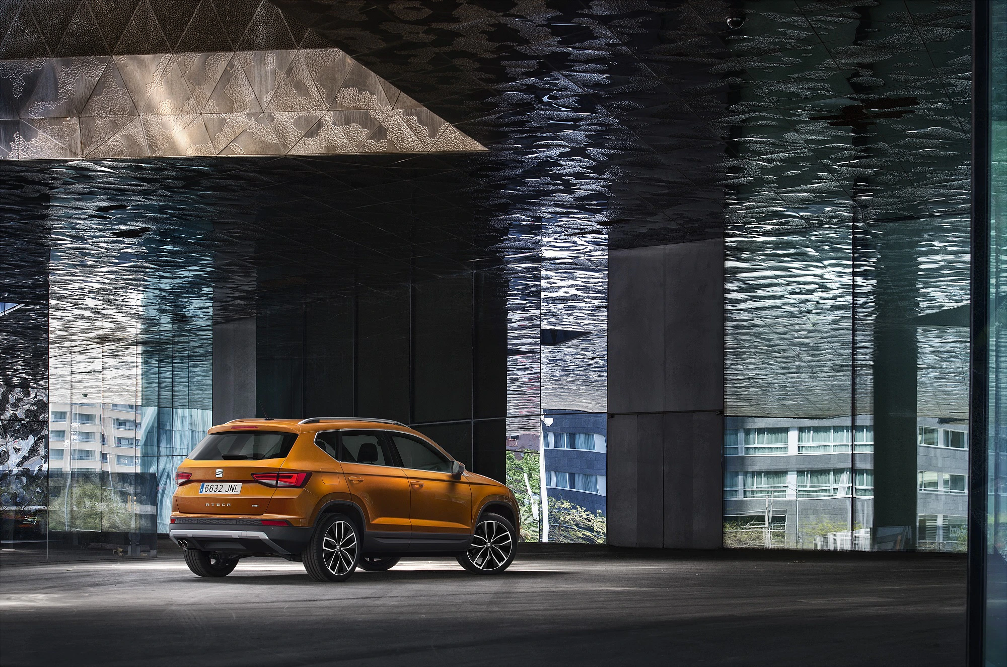 Orangefarbener SUV in modernem, düsterem Gebäude mit verspiegelter Decke und Glaswänden. Urbanes Ambiente mit Blick auf andere Gebäude.