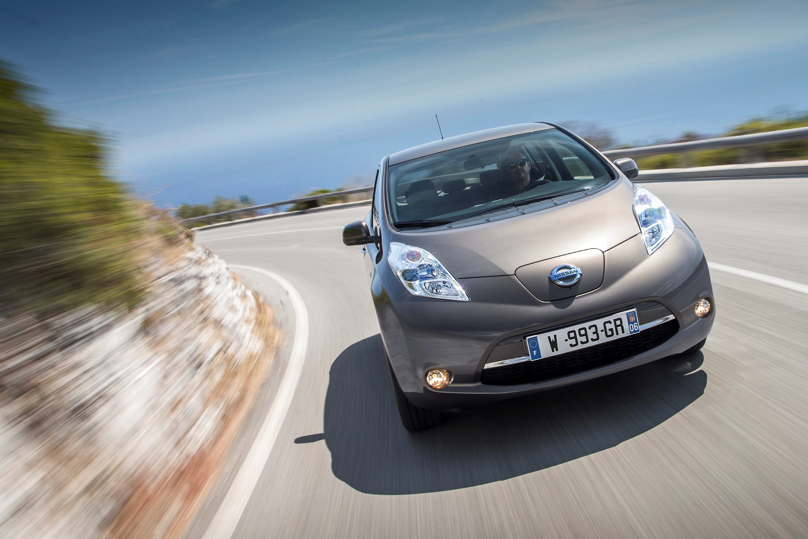 Silberner Nissan Leaf fährt dynamisch auf einer kurvigen Küstenstraße bei sonnigem Wetter.