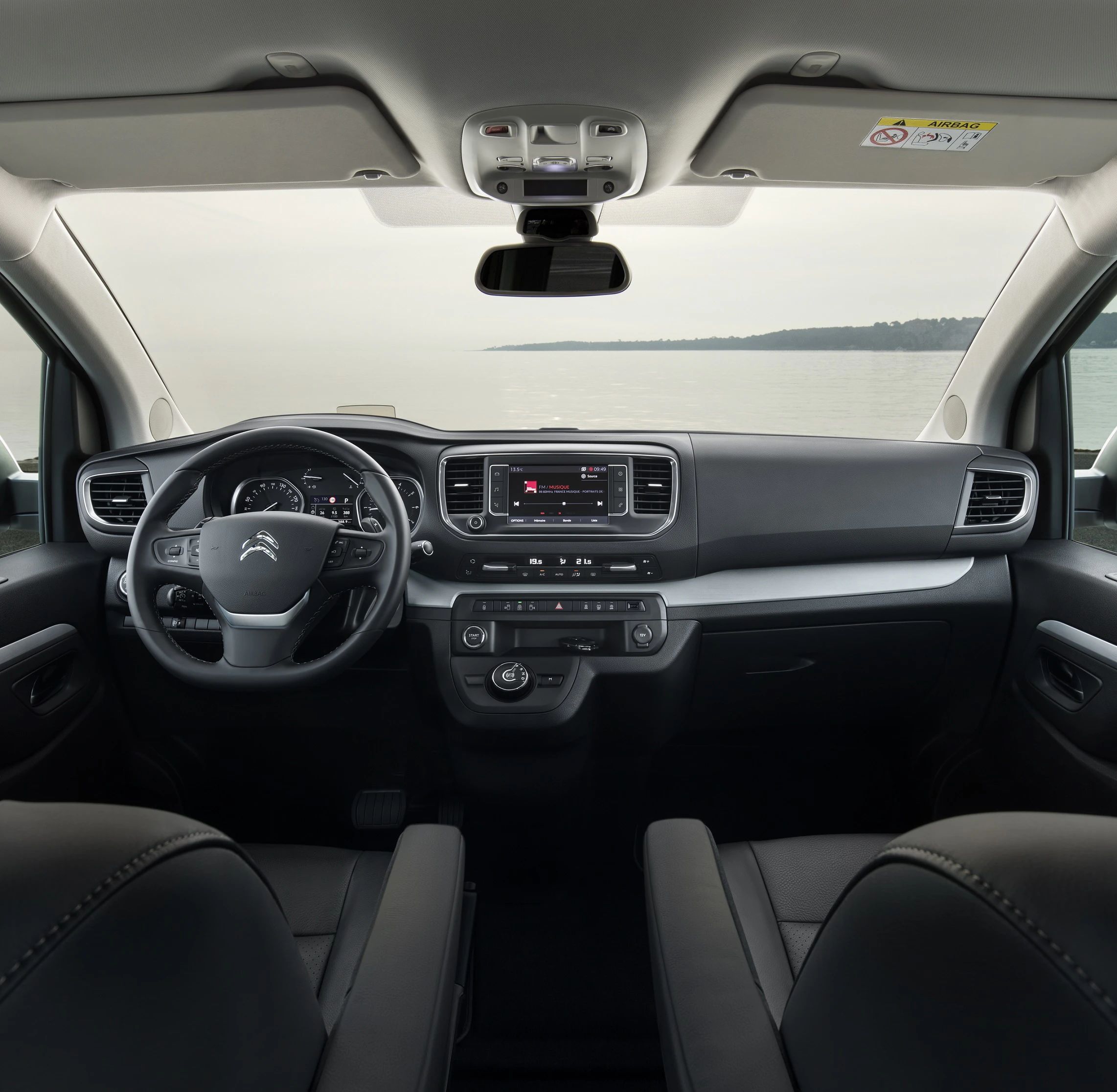 Innenraum eines modernen Autos mit Blick aufs Meer. Lenkrad links, Mittelkonsole mit Display und Bedienelementen, Ledersitze vorne.