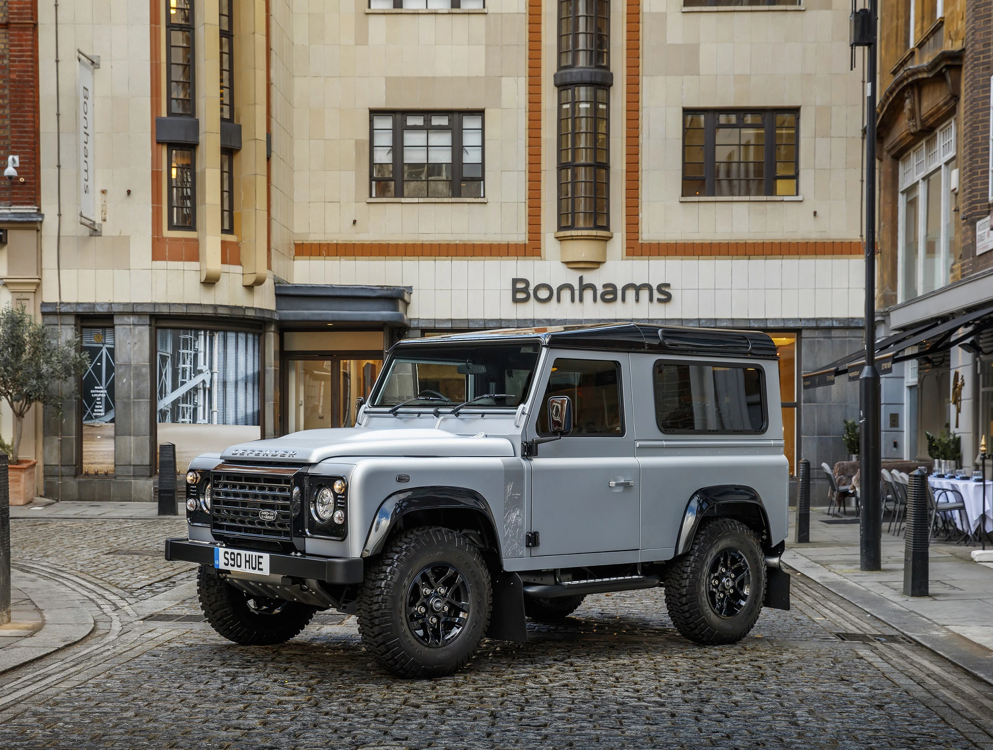 Silberner Geländewagen steht vor dem Bonhams-Gebäude in einer gepflasterten Straße, umgeben von urbaner Architektur.